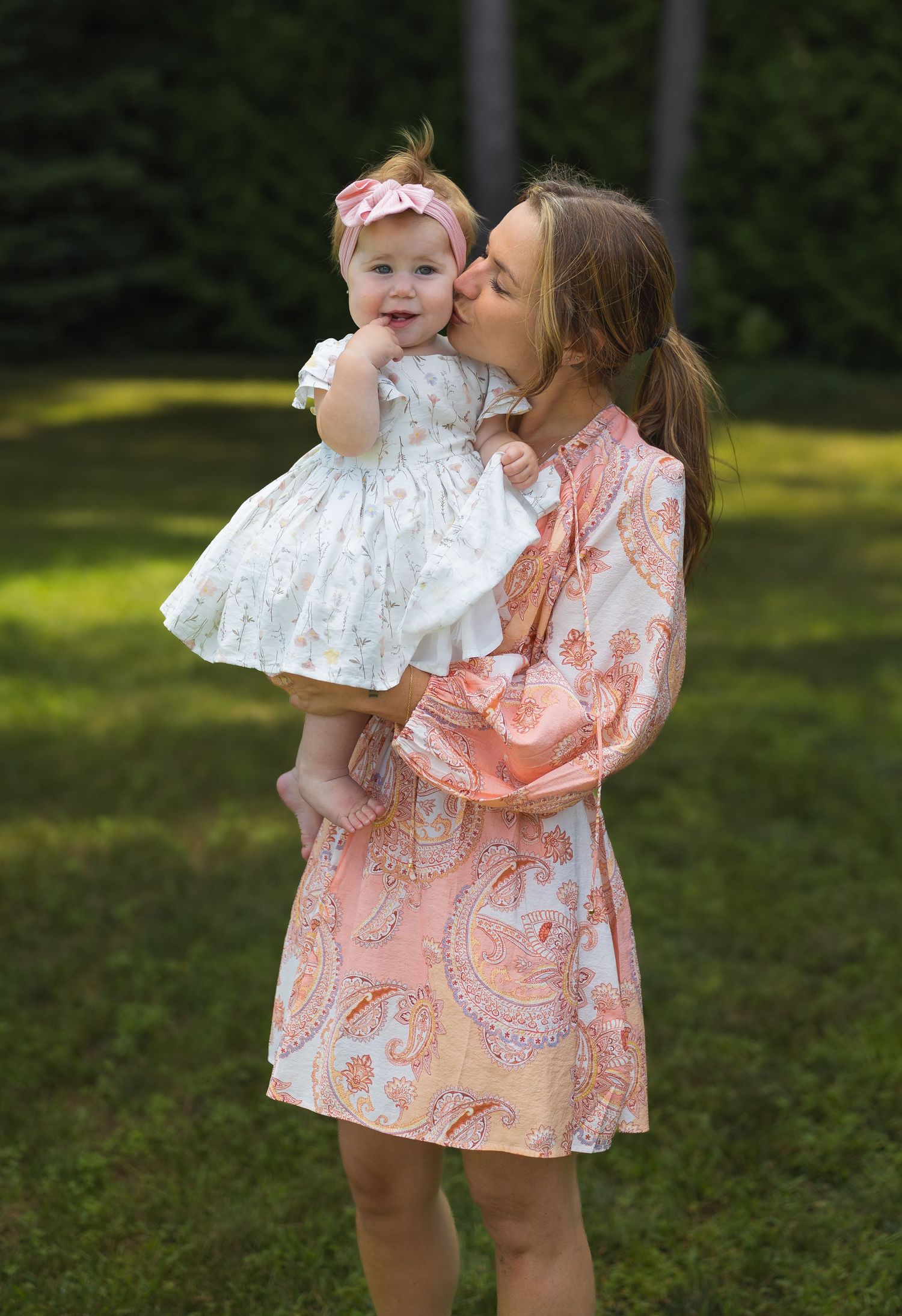 A mother in a peach paisley dress kisses her baby girl in a white dress with a pink bow headband outdoors.