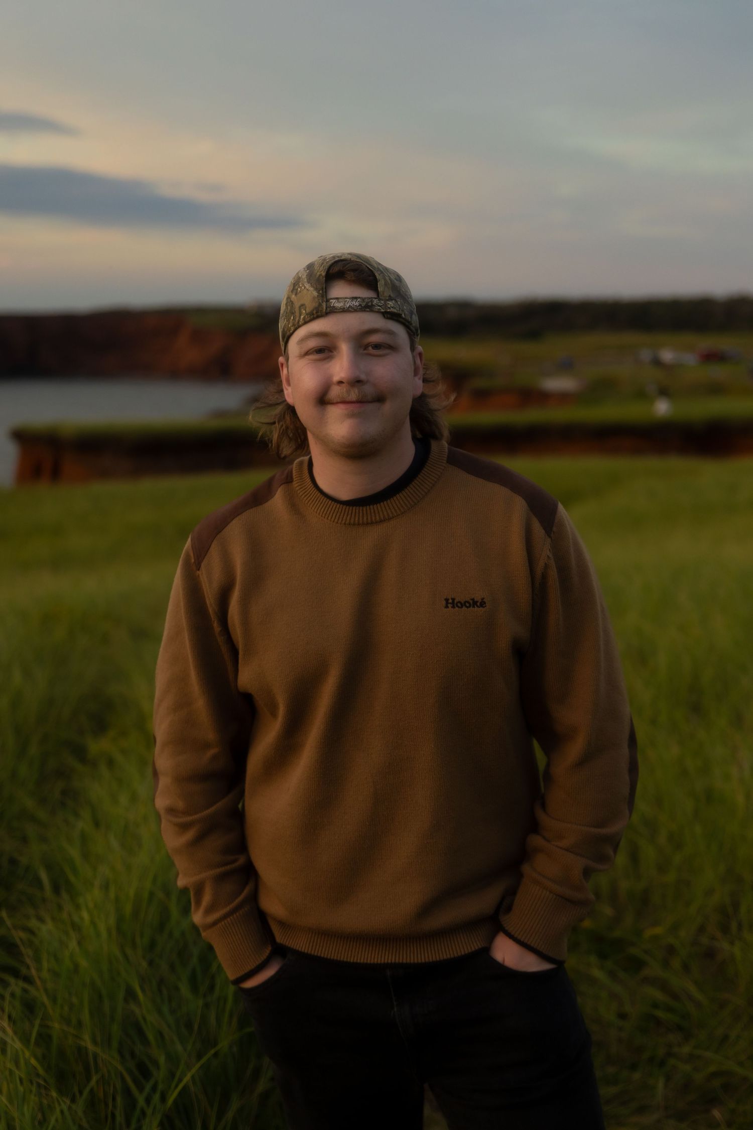 Young man in brown Hoodie sweatshirt and camo cap smiling outdoors at sunset near a green field and water.