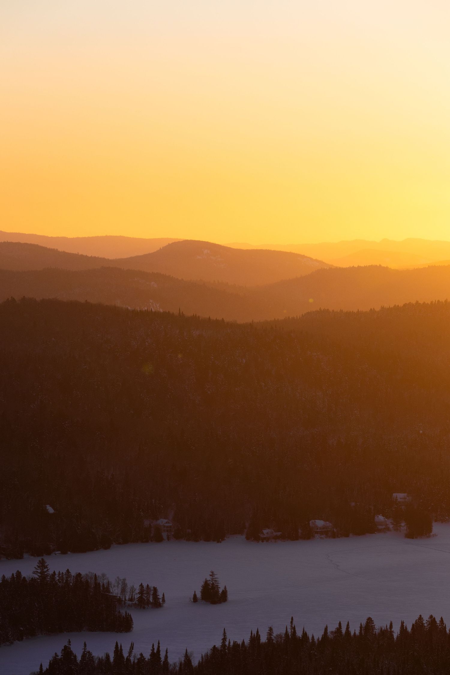 Orange sunset illuminates rolling mountain ridges with snow-covered forest and valley below.