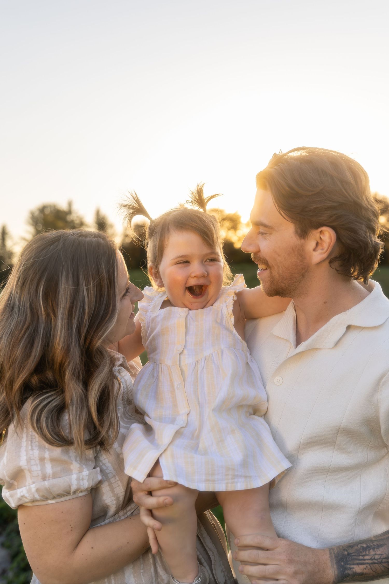 Family shares tender moments together in a grassy field at sunset wearing white and cream colored outfits.