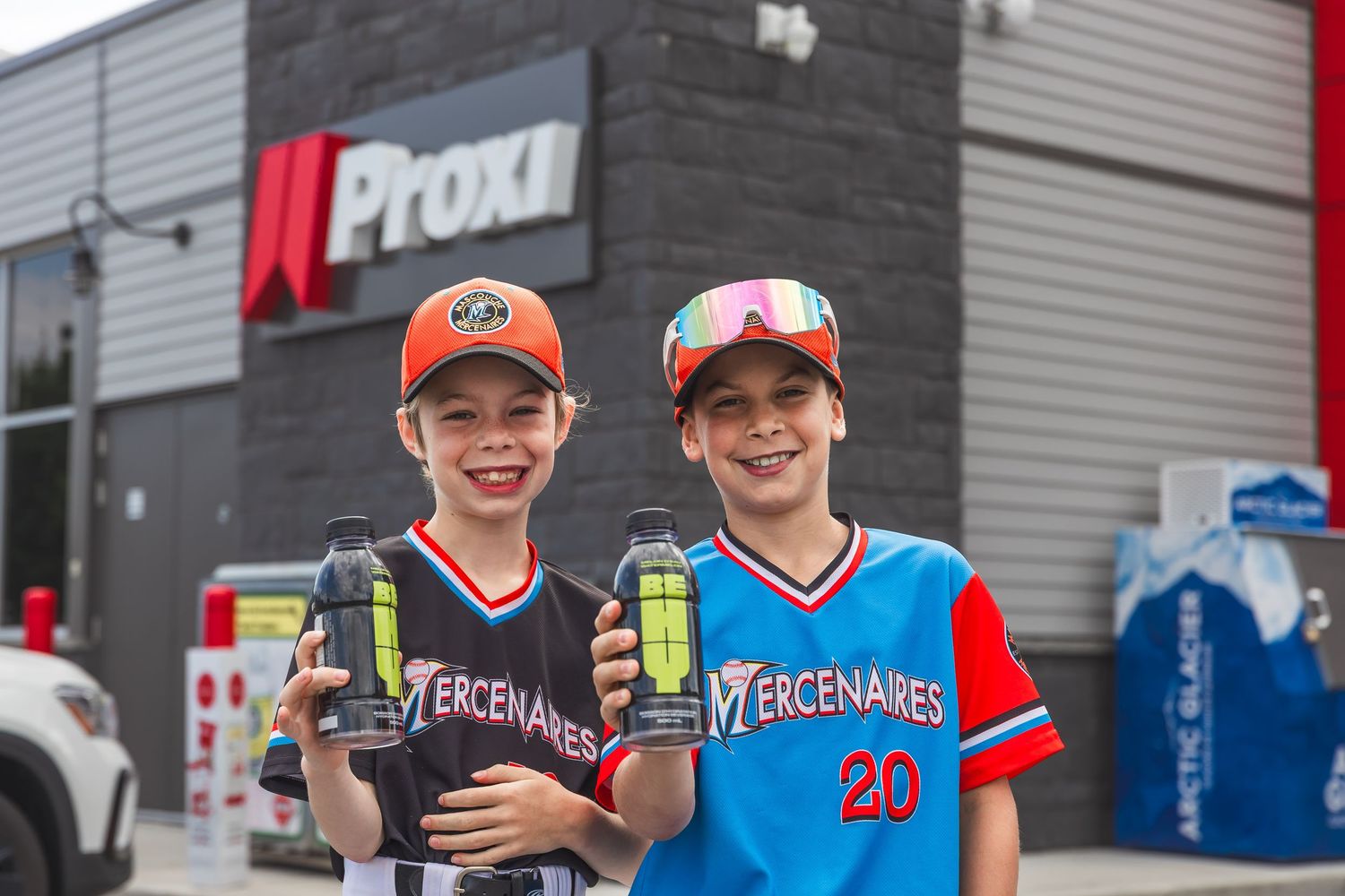 Youth baseball players in red and blue uniforms smile together during a game or practice session.