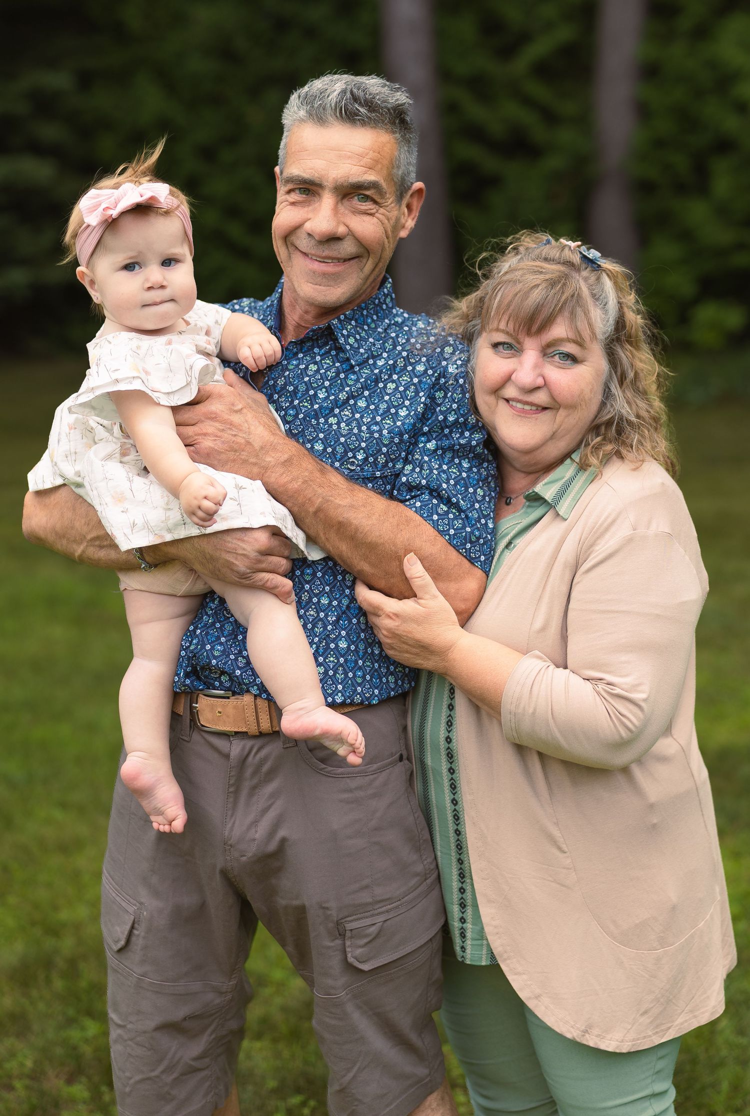 Smiling grandparents holding a cute baby girl in a white dress outdoors on a green lawn.