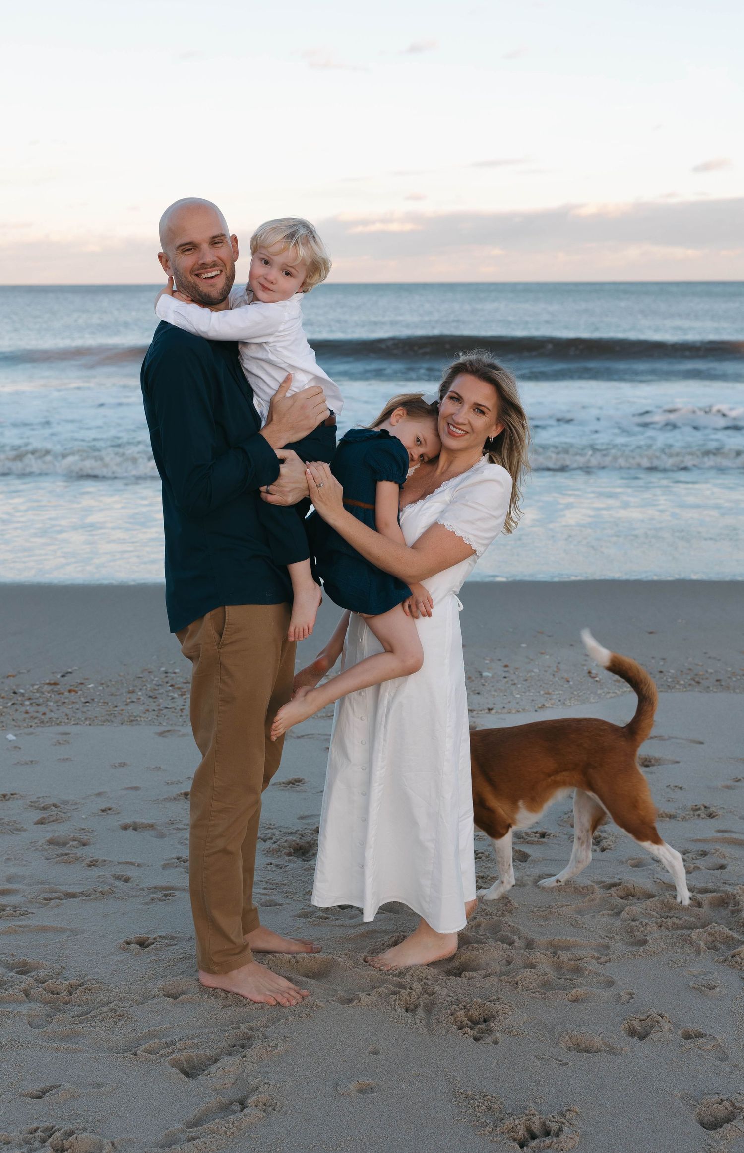Family enjoying a playful moment together on a sandy beach at sunset with their dog running alongside them.