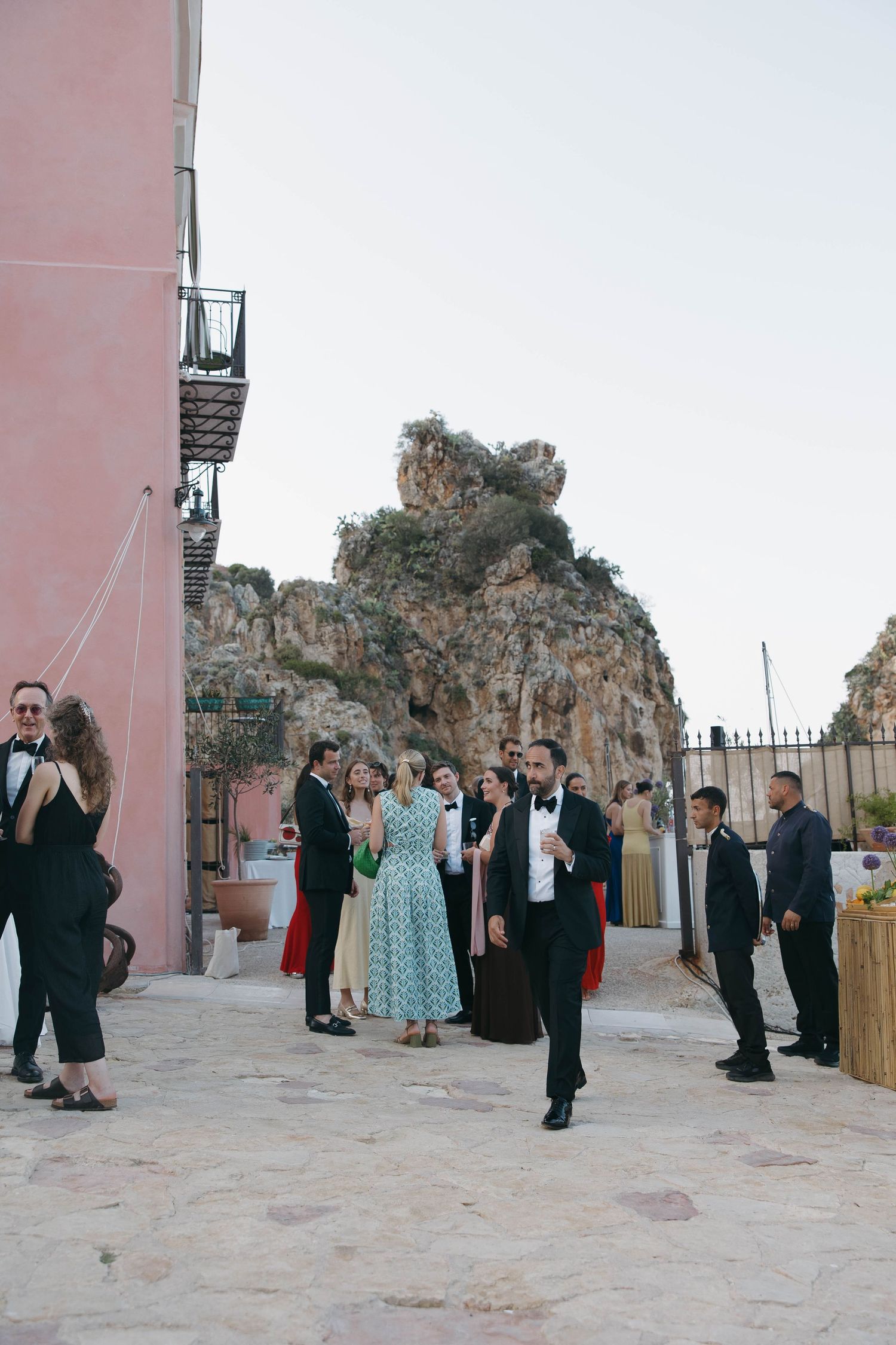 Wedding guests socialize in courtyard with pink building and coastal cliffs in background.