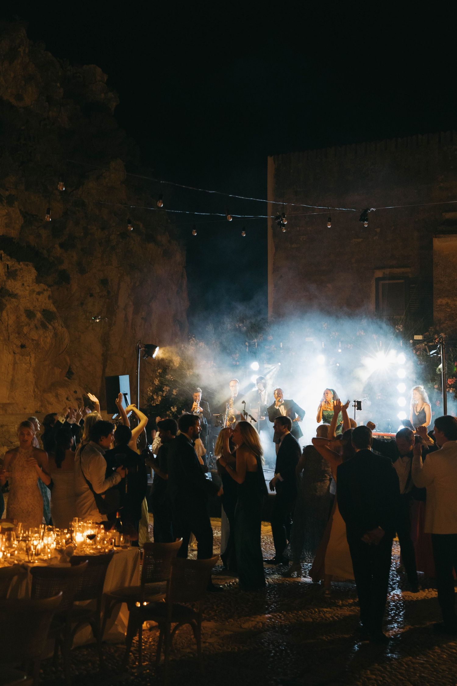 Nighttime outdoor celebration with crowd gathered around candlelit tables and atmospheric lighting.