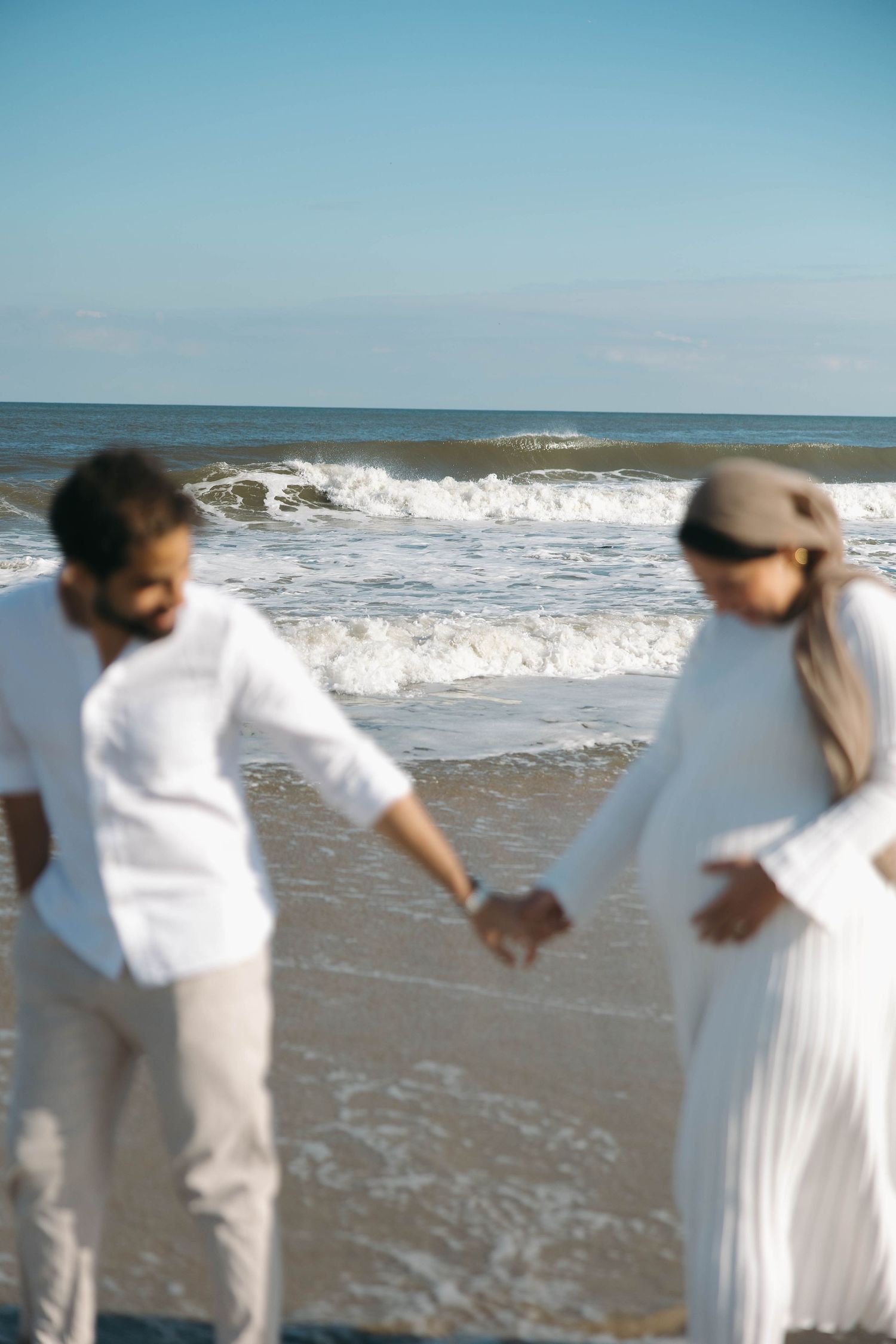 Two people in white outfits dance together on a sunny beach with ocean waves in the background.