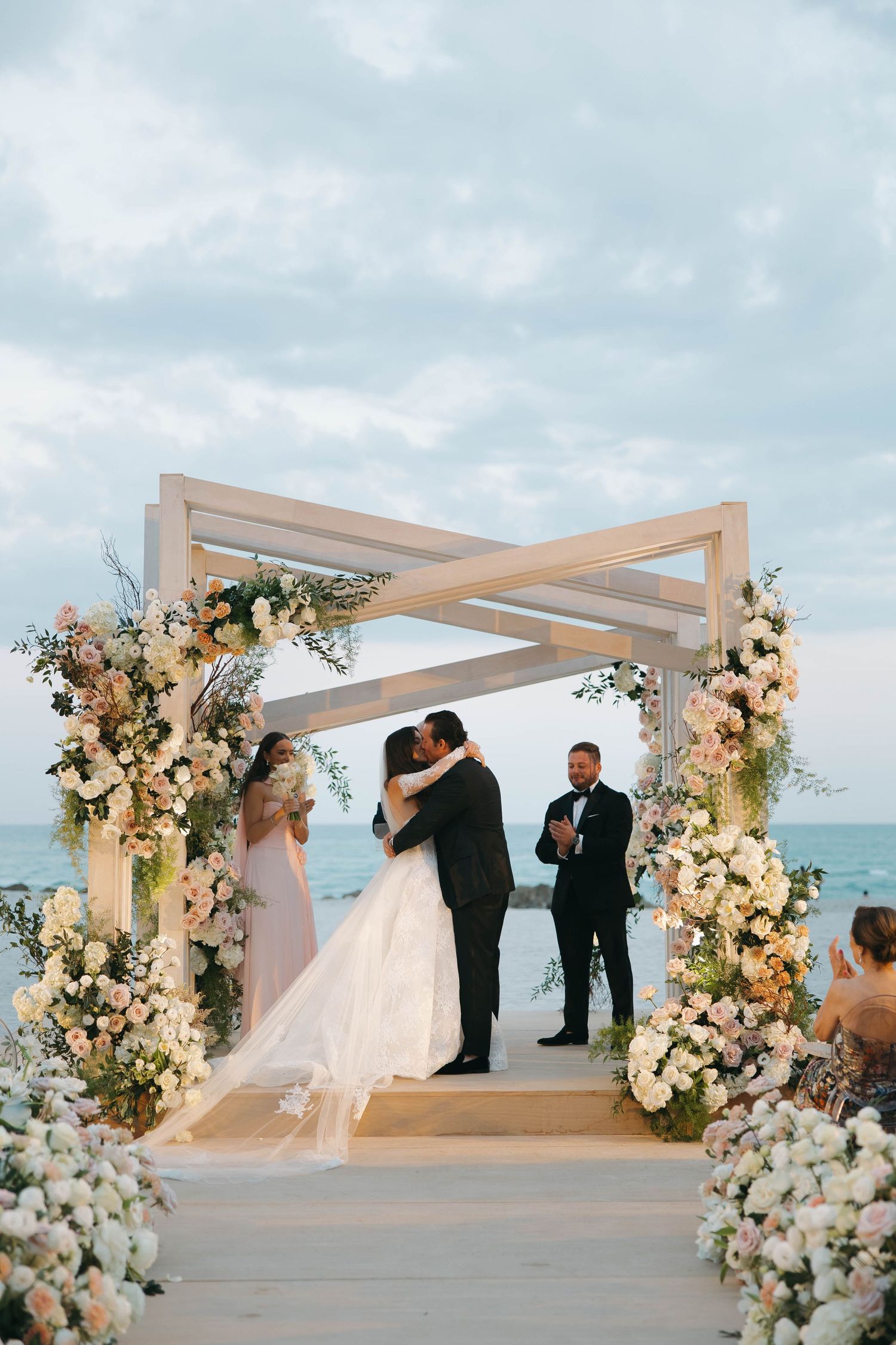 Romantic beach wedding ceremony taking place under an elegant white pergola with floral arrangements.