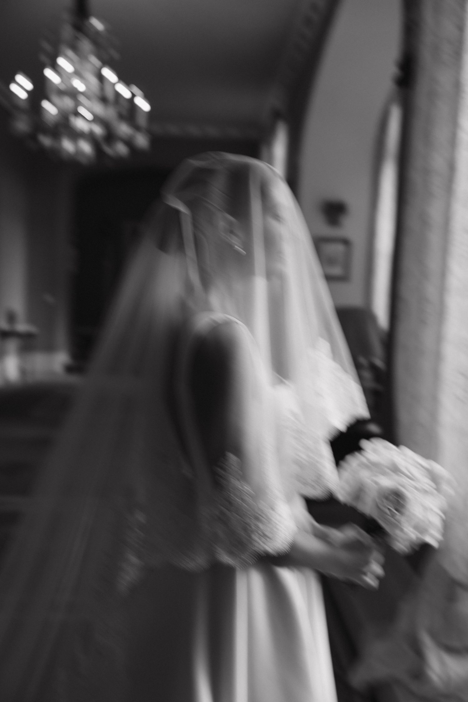 Artistic black and white photo of bride in flowing veil holding bouquet indoors.