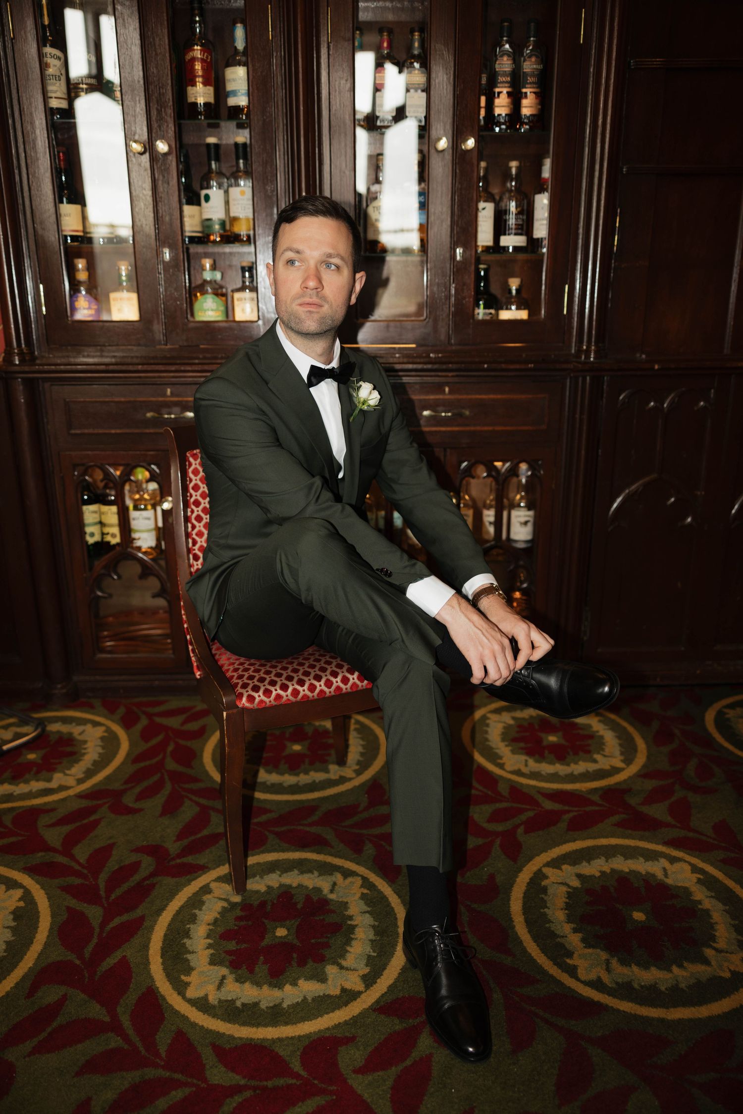 A groomsman in a black tuxedo sits on an ornate carpet in front of a wooden bar with liquor bottles.