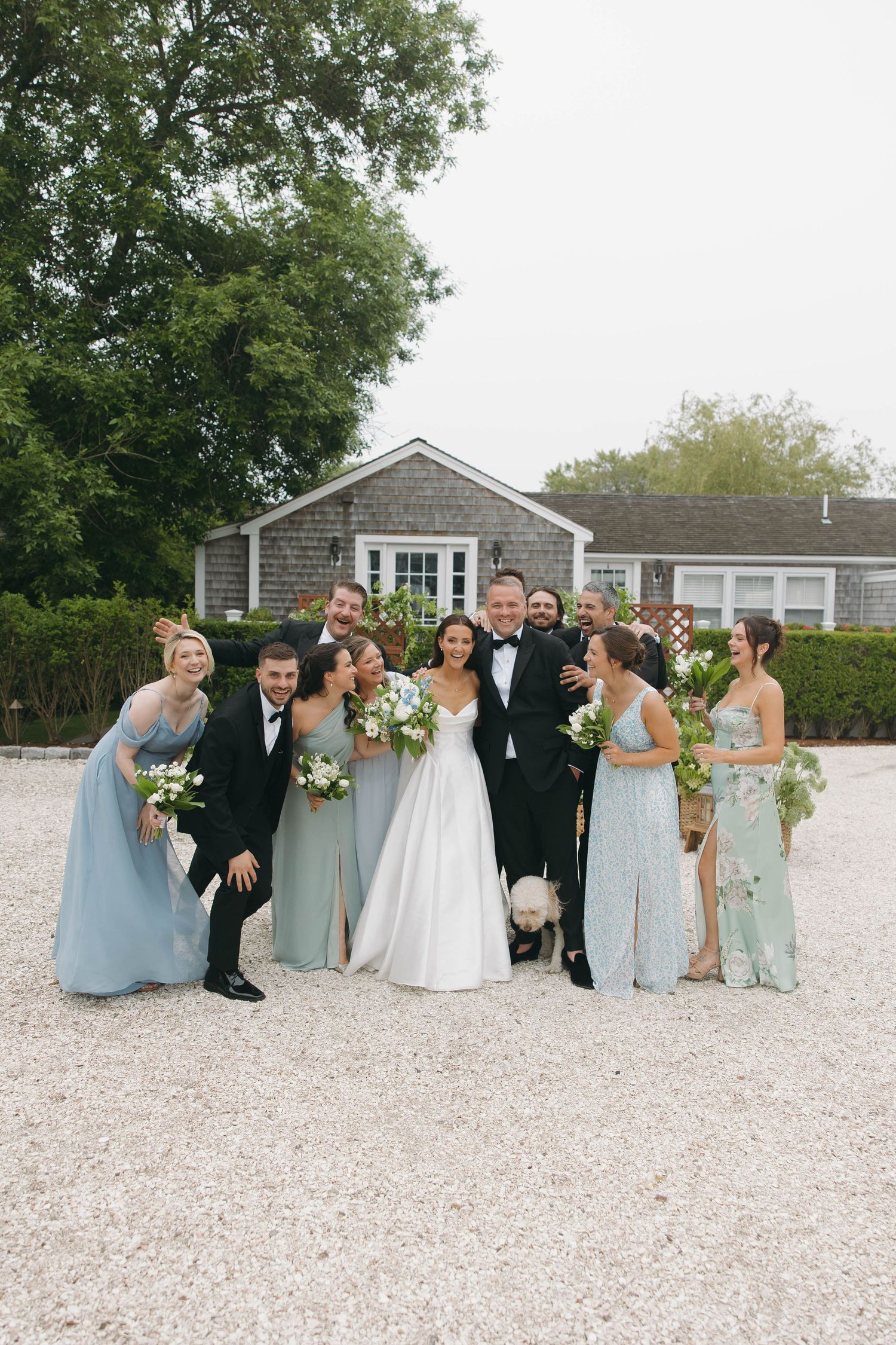 Wedding party members pose and laugh together on gravel patio in front of grey shingled building.