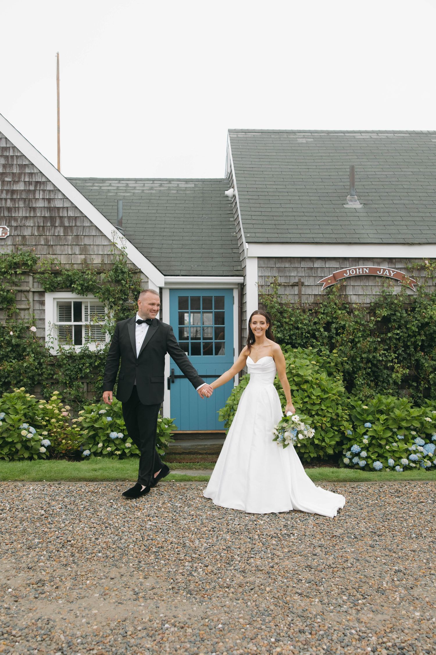 A couple in wedding attire poses playfully outside a charming grey shingled cottage with blue door and hydrangea bushes.