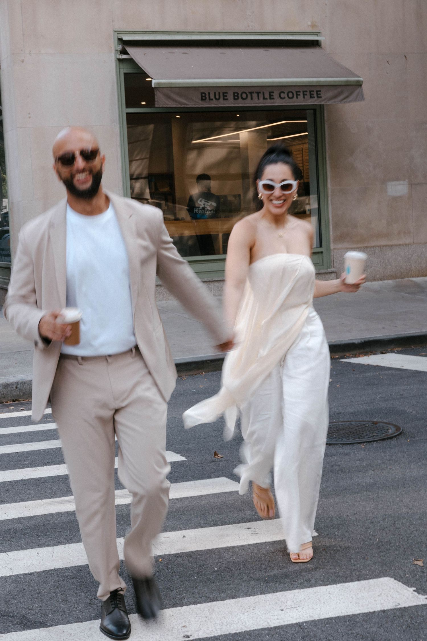 A couple in light colored outfits dances playfully on a crosswalk near Blue Bottle Coffee.