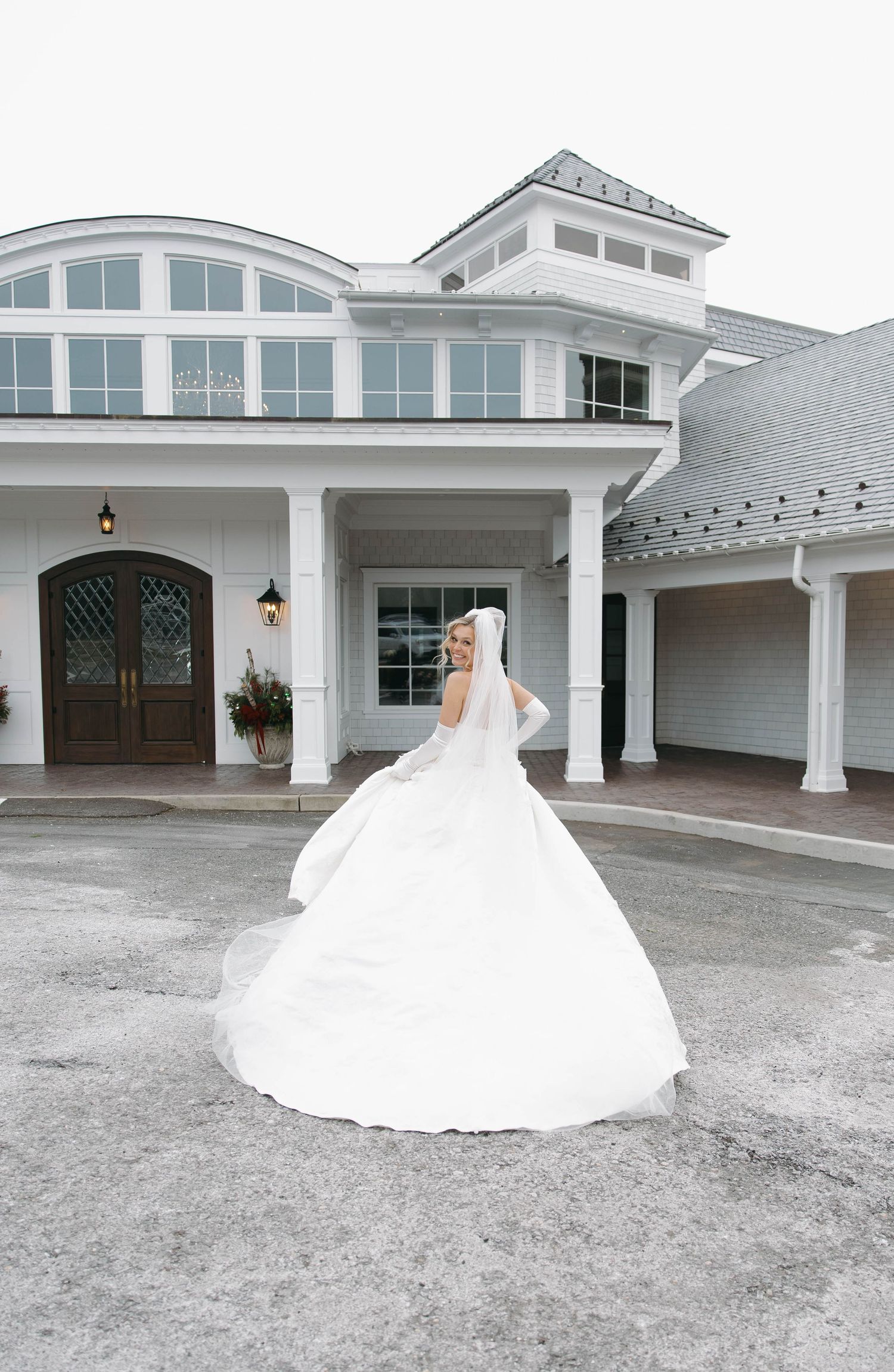 A bride in a dramatic ballgown poses outside a white coastal wedding venue.