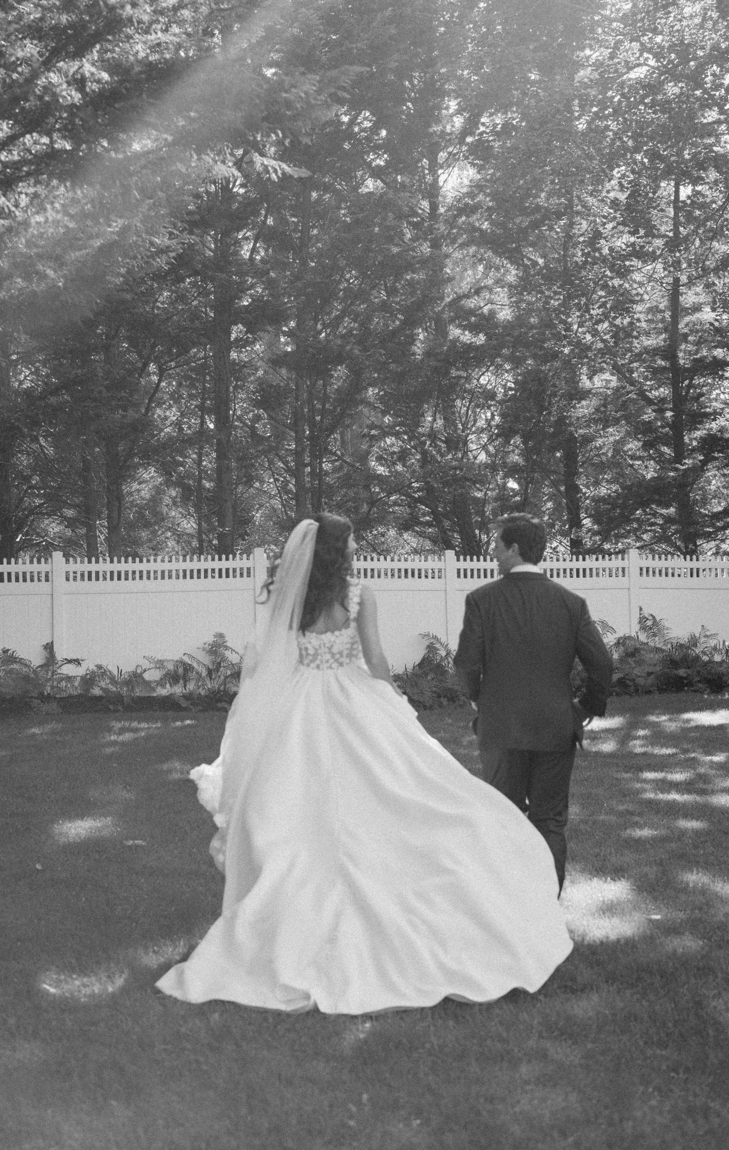 A couple walks across a sunlit lawn with white picket fence in the background.
