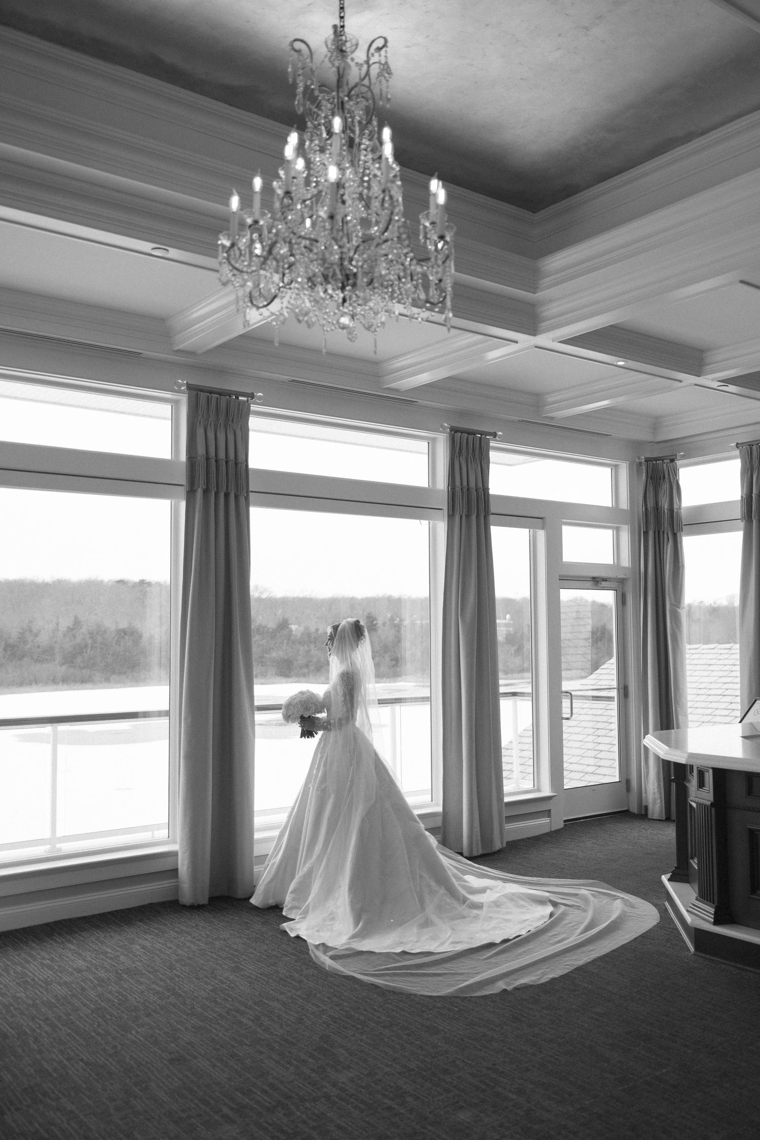 A wedding dress displayed in an elegant room with large windows, chandeliers and gray curtains.