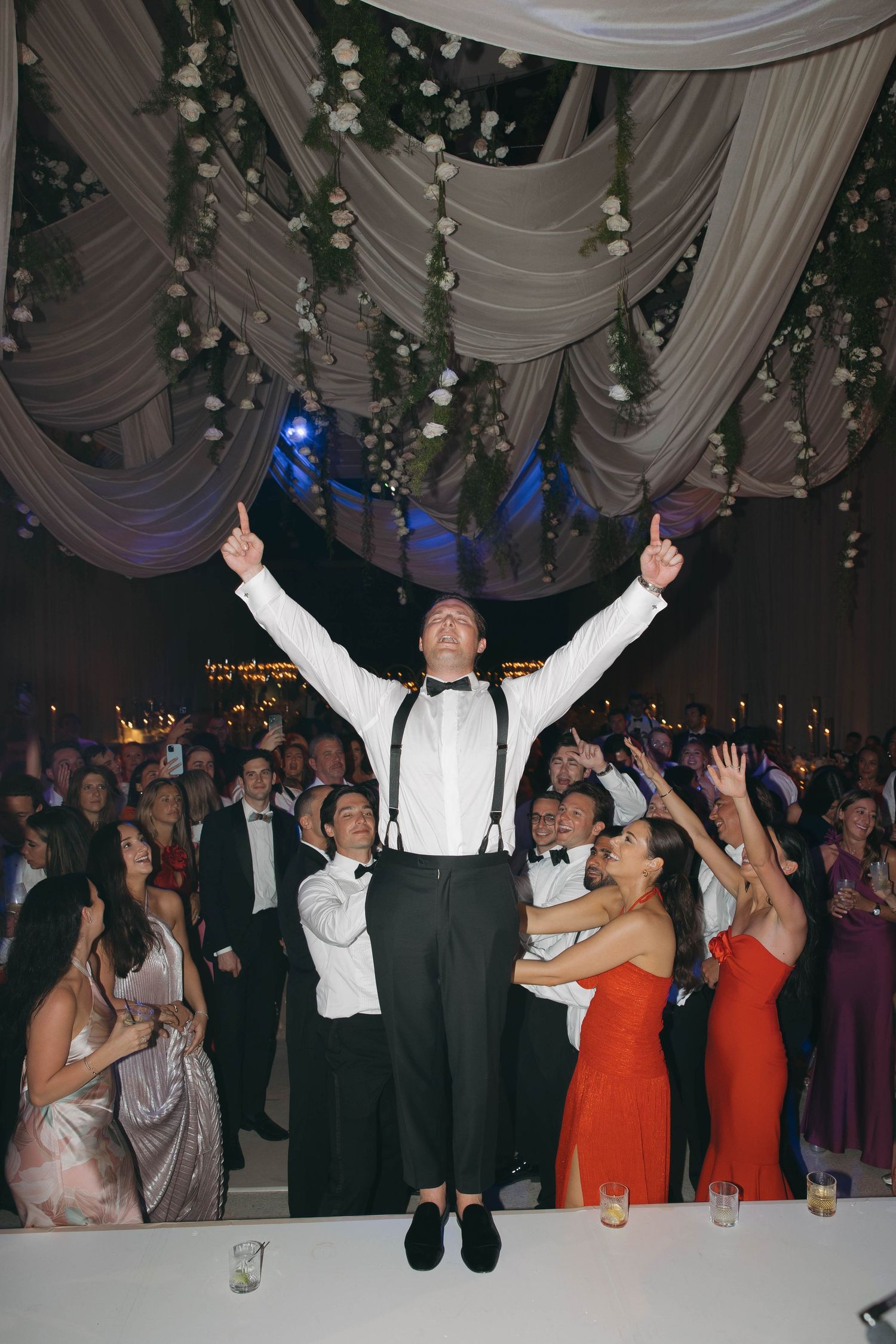 Wedding celebration with guests jumping and dancing under decorative ceiling draping.