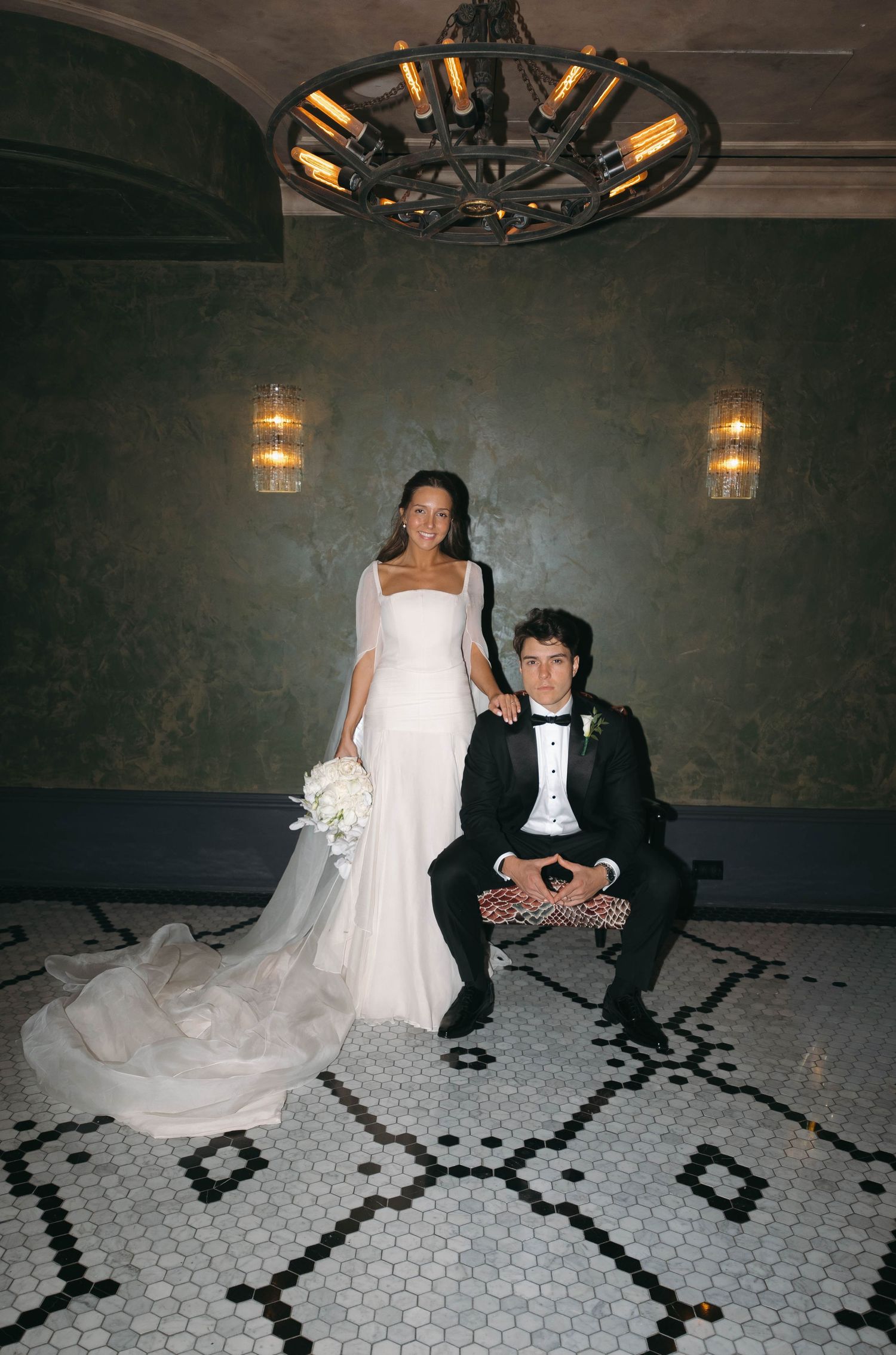 Bride in sleek white gown with veil stands beside seated groom in tuxedo under chandelier on mosaic tile floor.