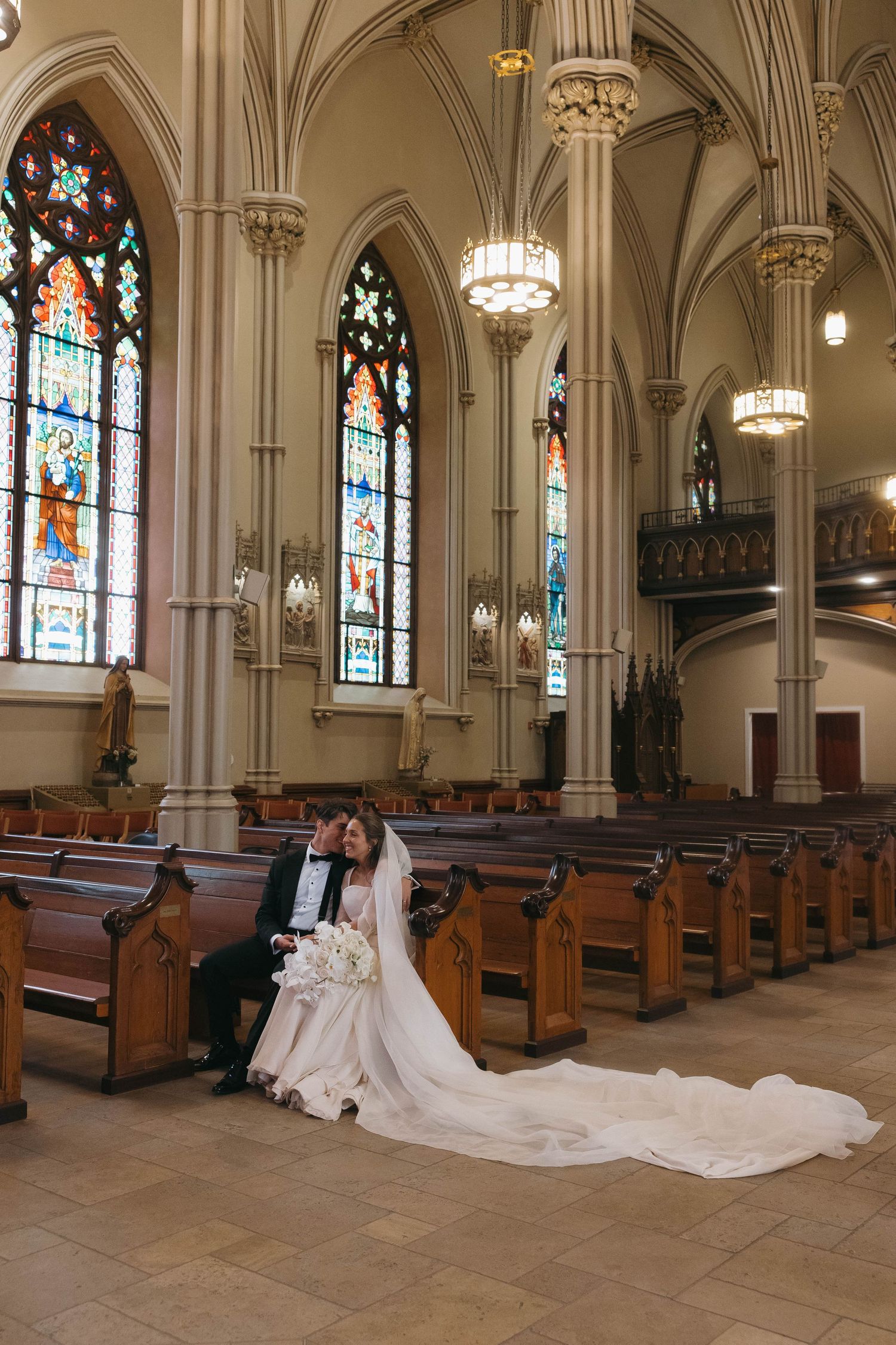 Bride and groom sit together on church pew surrounded by Gothic arches and colorful stained glass windows.