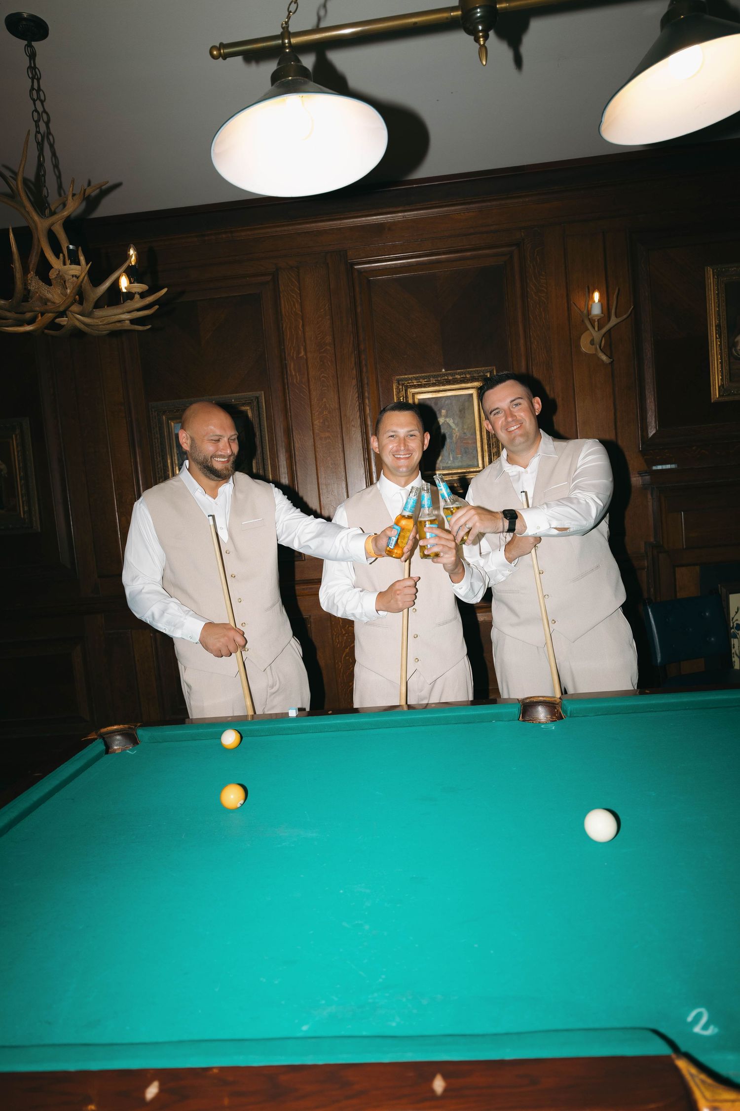 Three groomsmen in beige suits celebrating around a pool table in a wood-paneled billiard room.