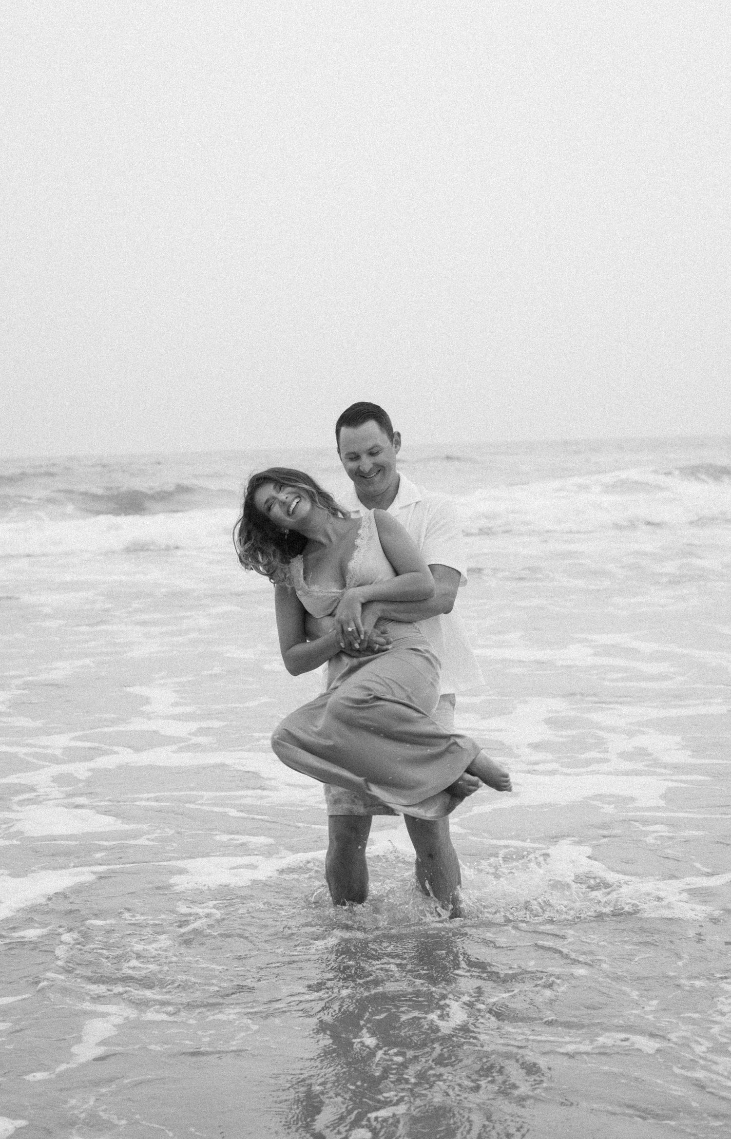 A couple plays and laughs together in ocean waves at the beach in black and white.