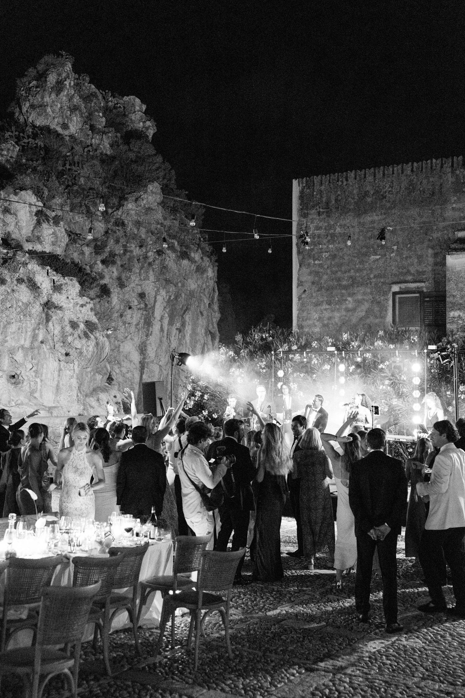 Black and white photo of an elegant outdoor reception with stone walls and string lights overhead.