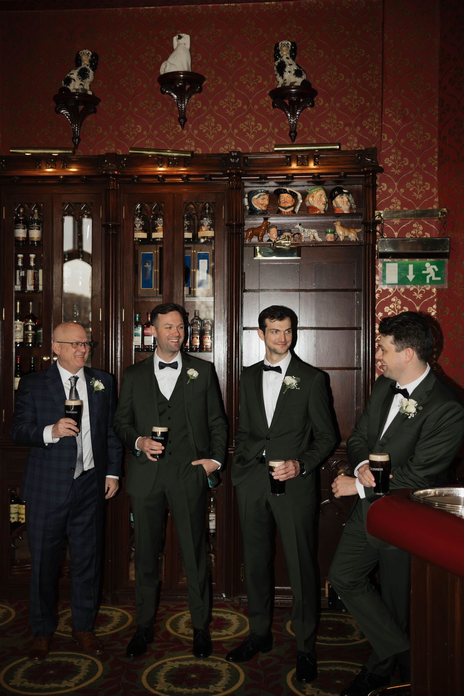 Wedding party members in black tuxedos gather at a bar with dark wood paneling and red wallpaper.