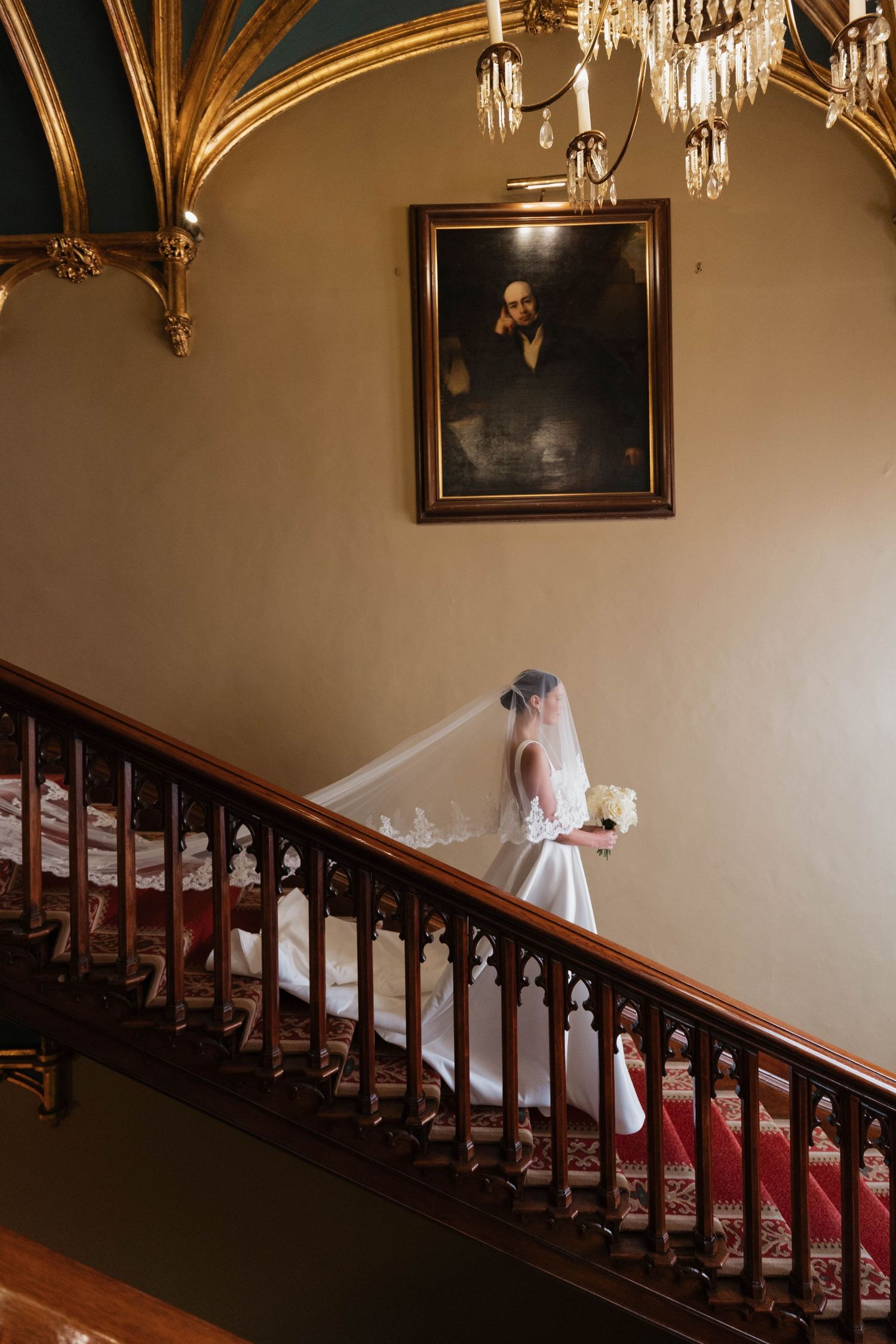 Bride in white gown walking down ornate staircase under crystal chandelier in warm lighting.