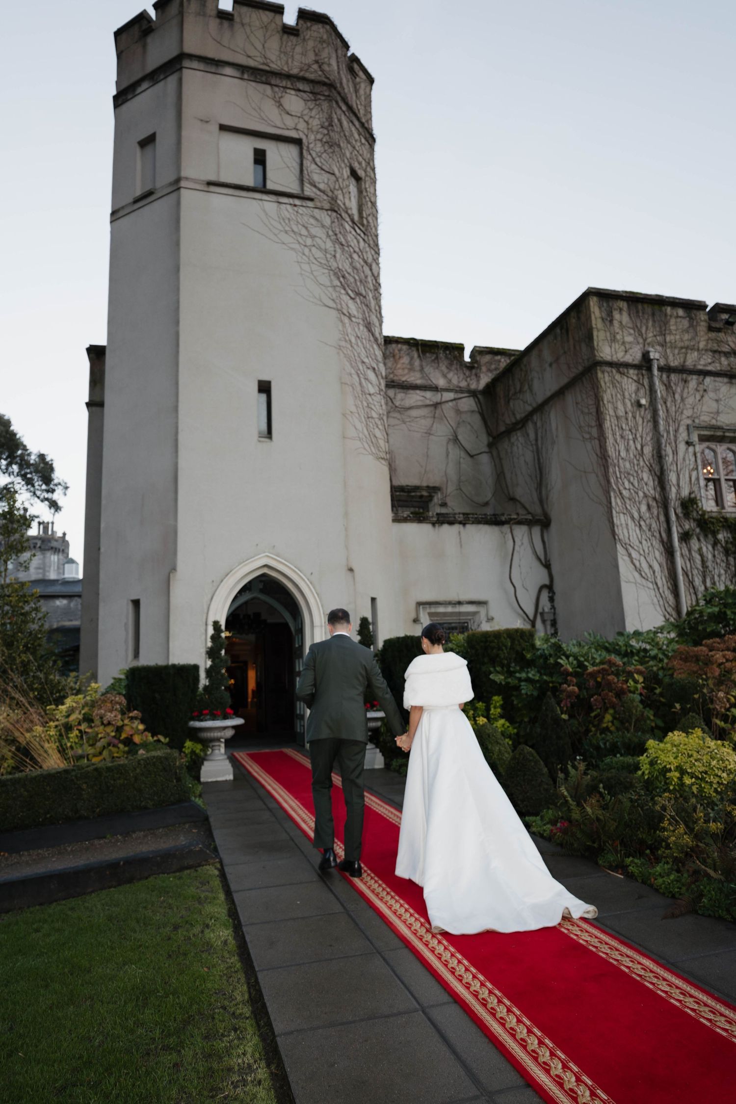 Wedding party entering castle-like venue on red carpet with white stone tower in background.