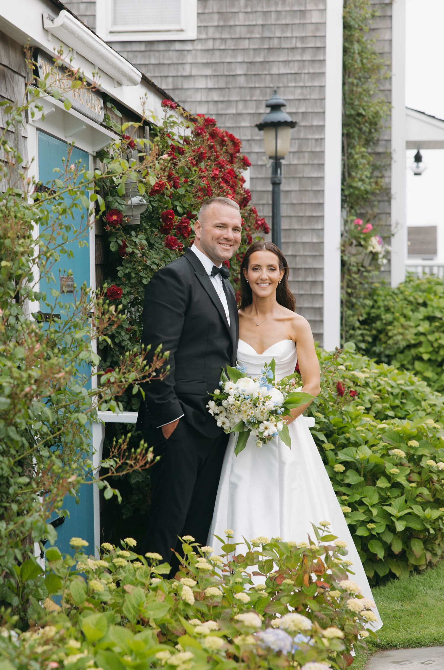 Elegant wedding portraits captured in front of a coastal cottage with blooming summer flowers.