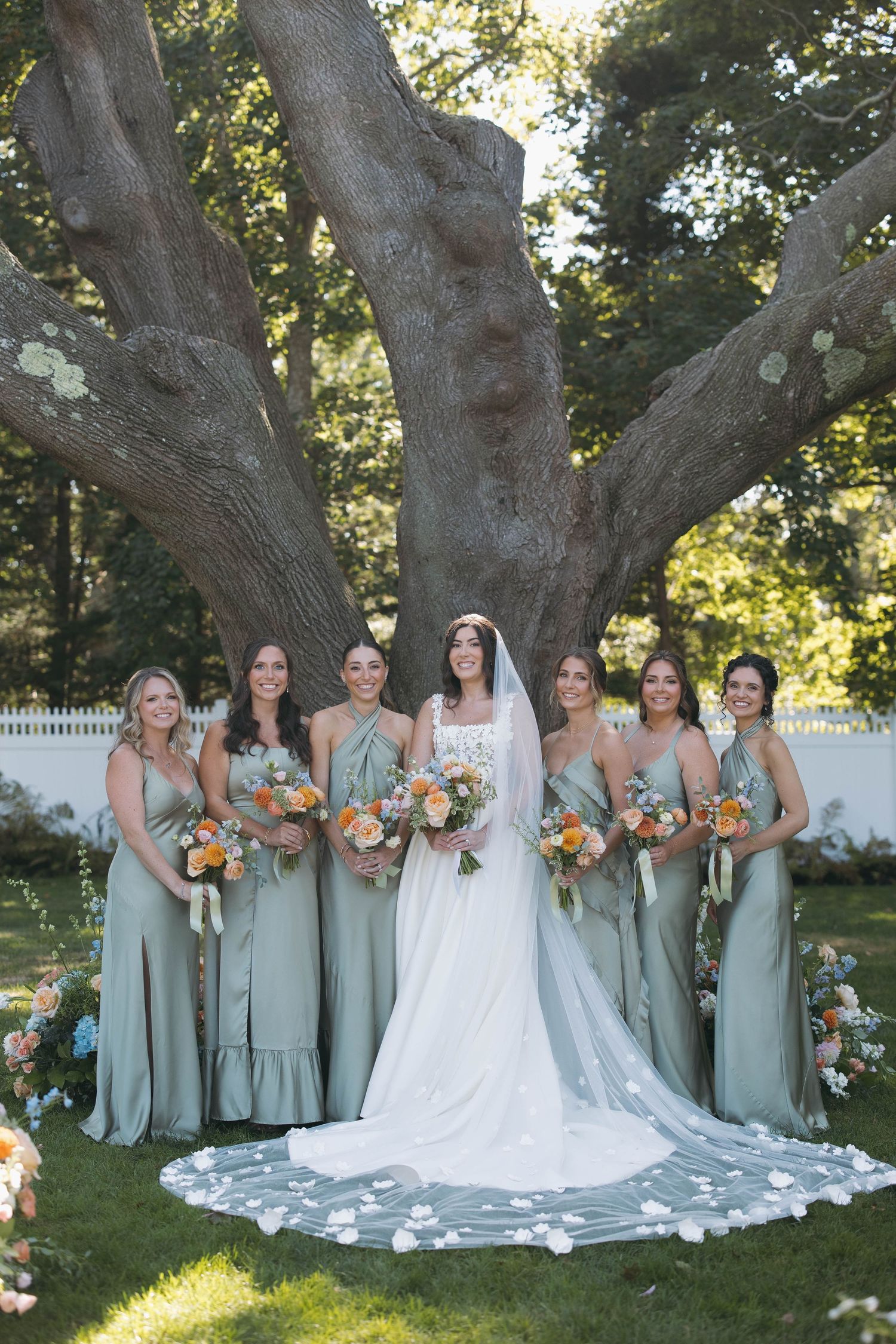 Bridal party in sage green dresses poses under a large tree with white fence in background.