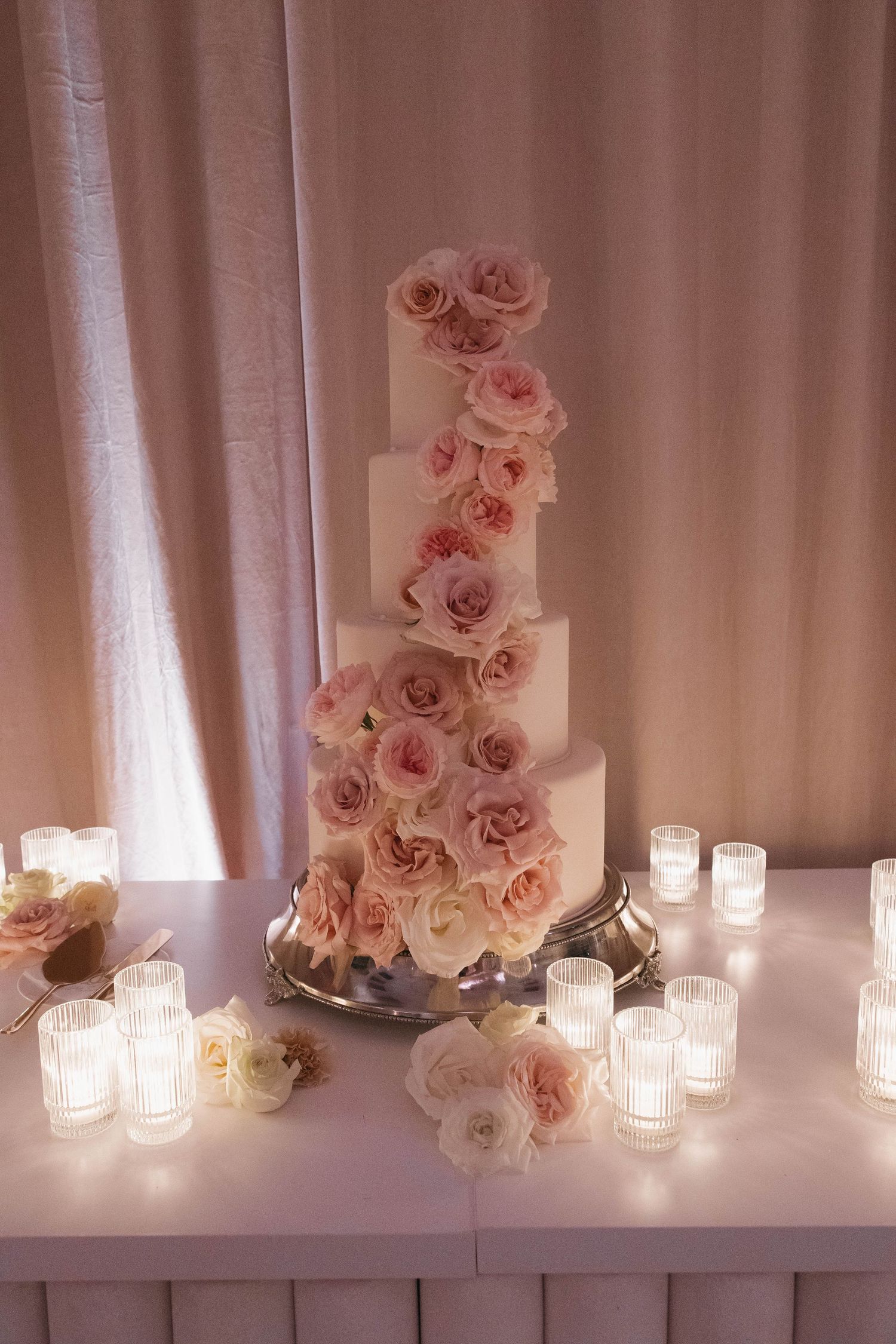 Pink roses and glowing candles arranged along white draped tables for wedding decor.