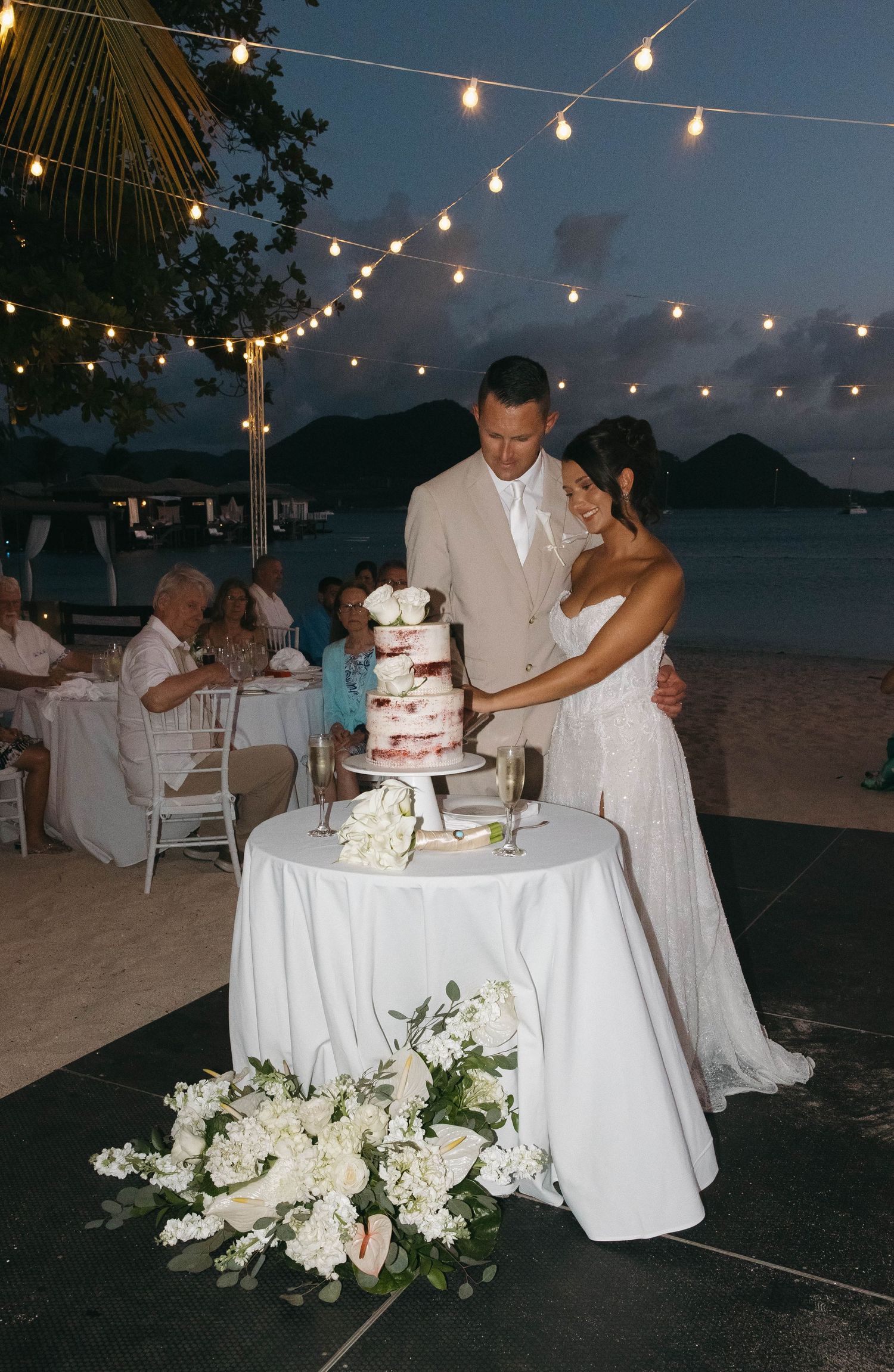 Bride and groom cutting wedding cake on beach at dusk with string lights and floral arrangements.