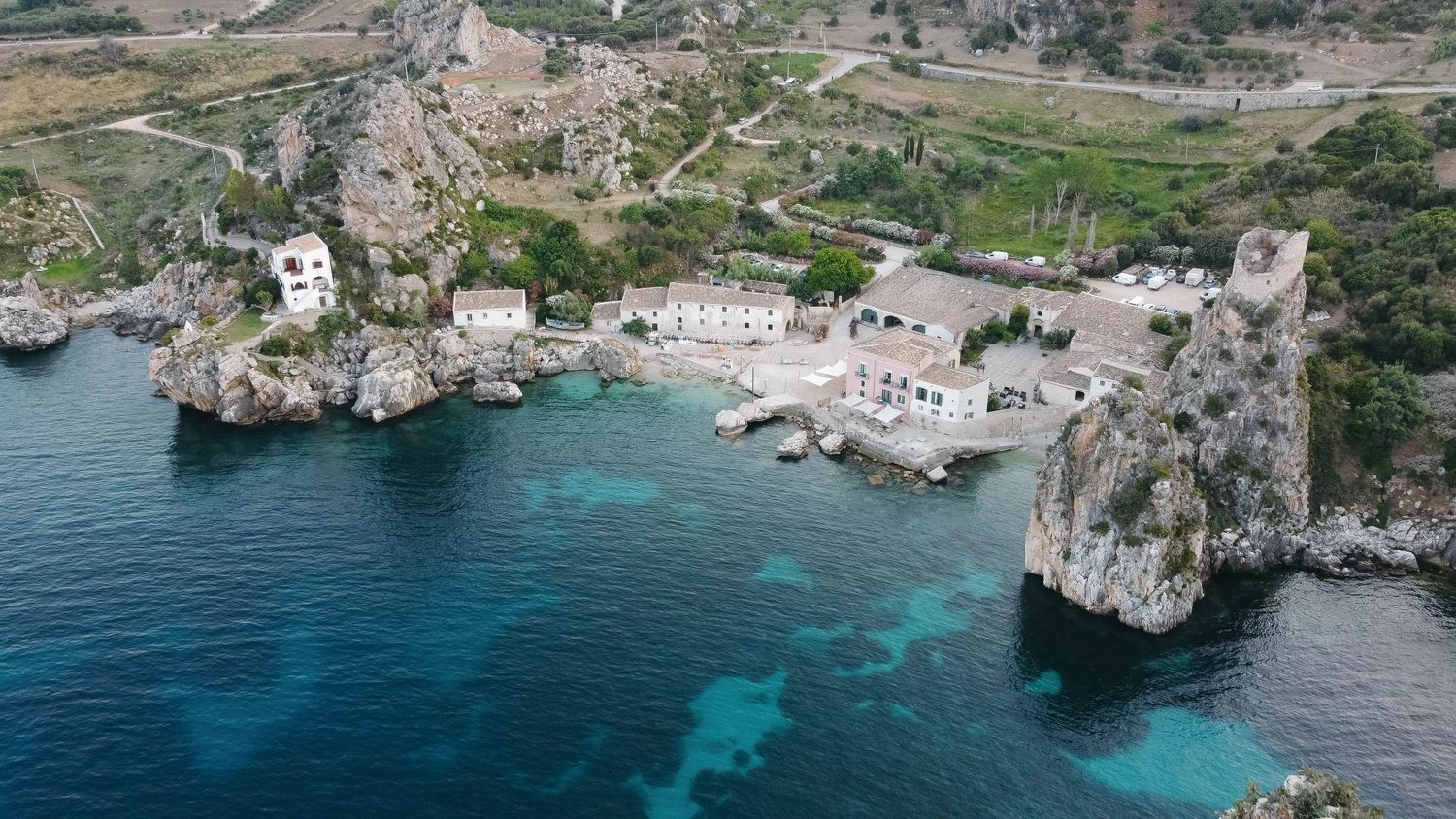 Sweeping aerial perspective of a Mediterranean bay with historic buildings and crystal clear waters.