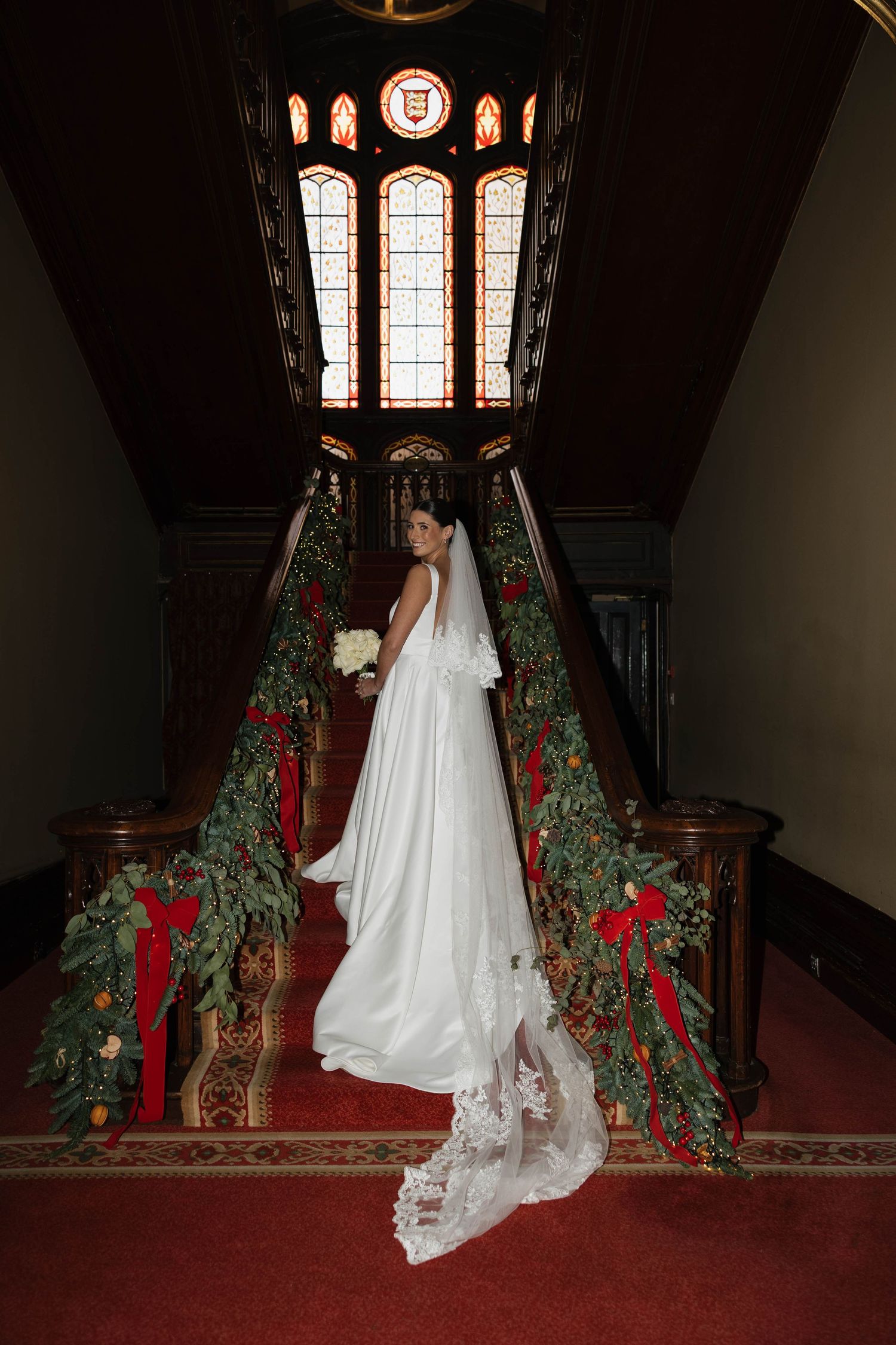Elegant white wedding dress with long lace train on red carpeted staircase decorated with greenery and red ribbons.