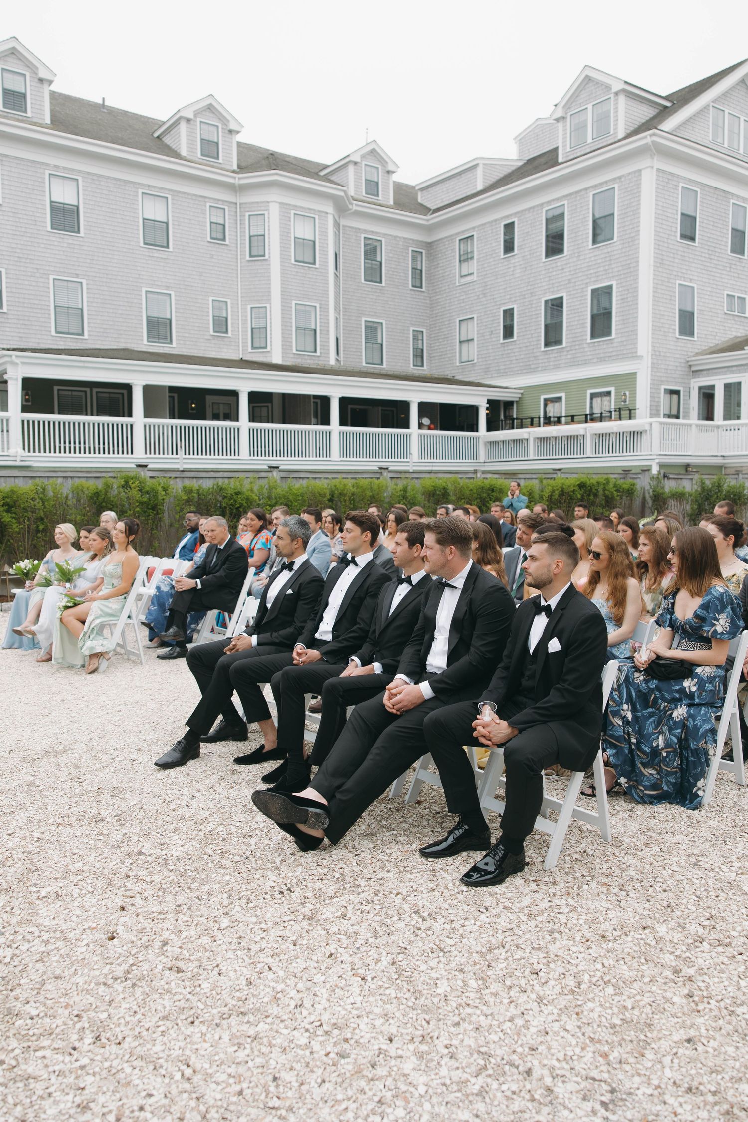 Wedding guests seated on white chairs with large coastal-style building in background.