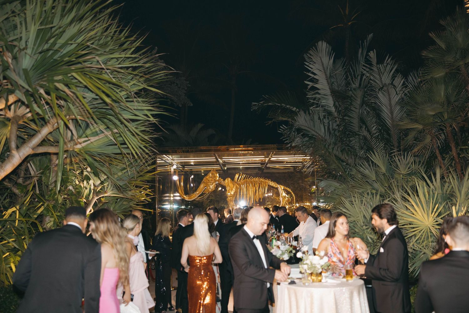 Nighttime outdoor reception with guests dining among tropical palm trees.