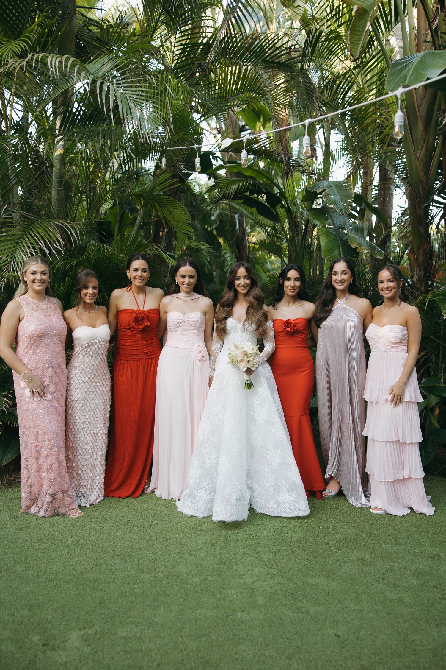 Bridesmaids in pink and red dresses pose together on green lawn with tropical backdrop.