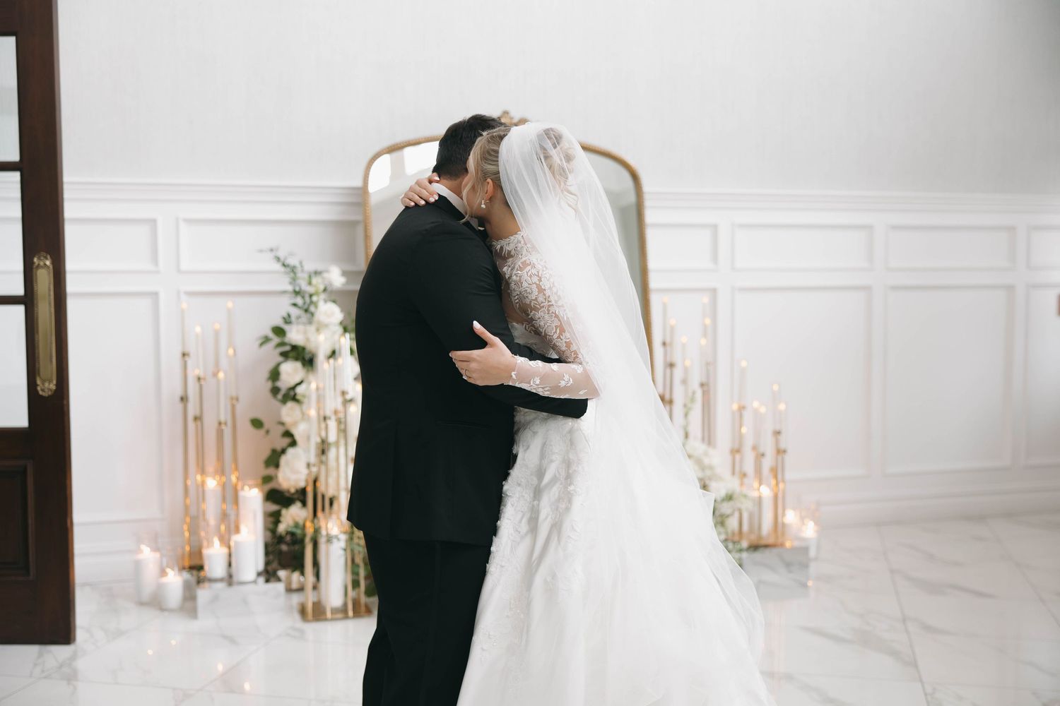 Wedding couple embracing in front of decorative mirror and floral arrangements.