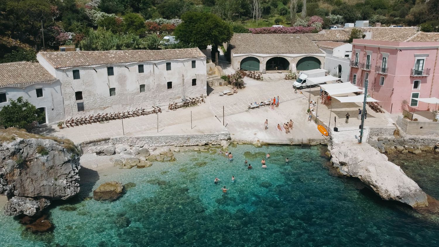 Aerial panorama of a Mediterranean coastal village with pink buildings and turquoise waters along rocky cliffs.
