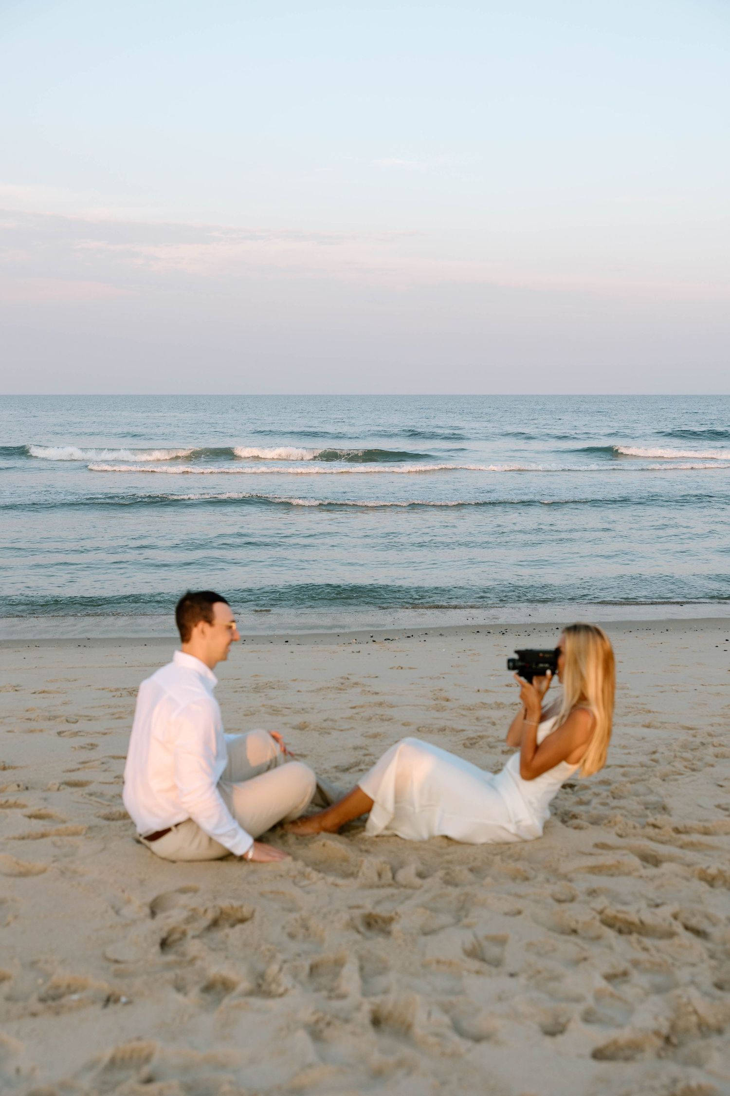 A couple lying on beach sand taking photos of each other at dusk.