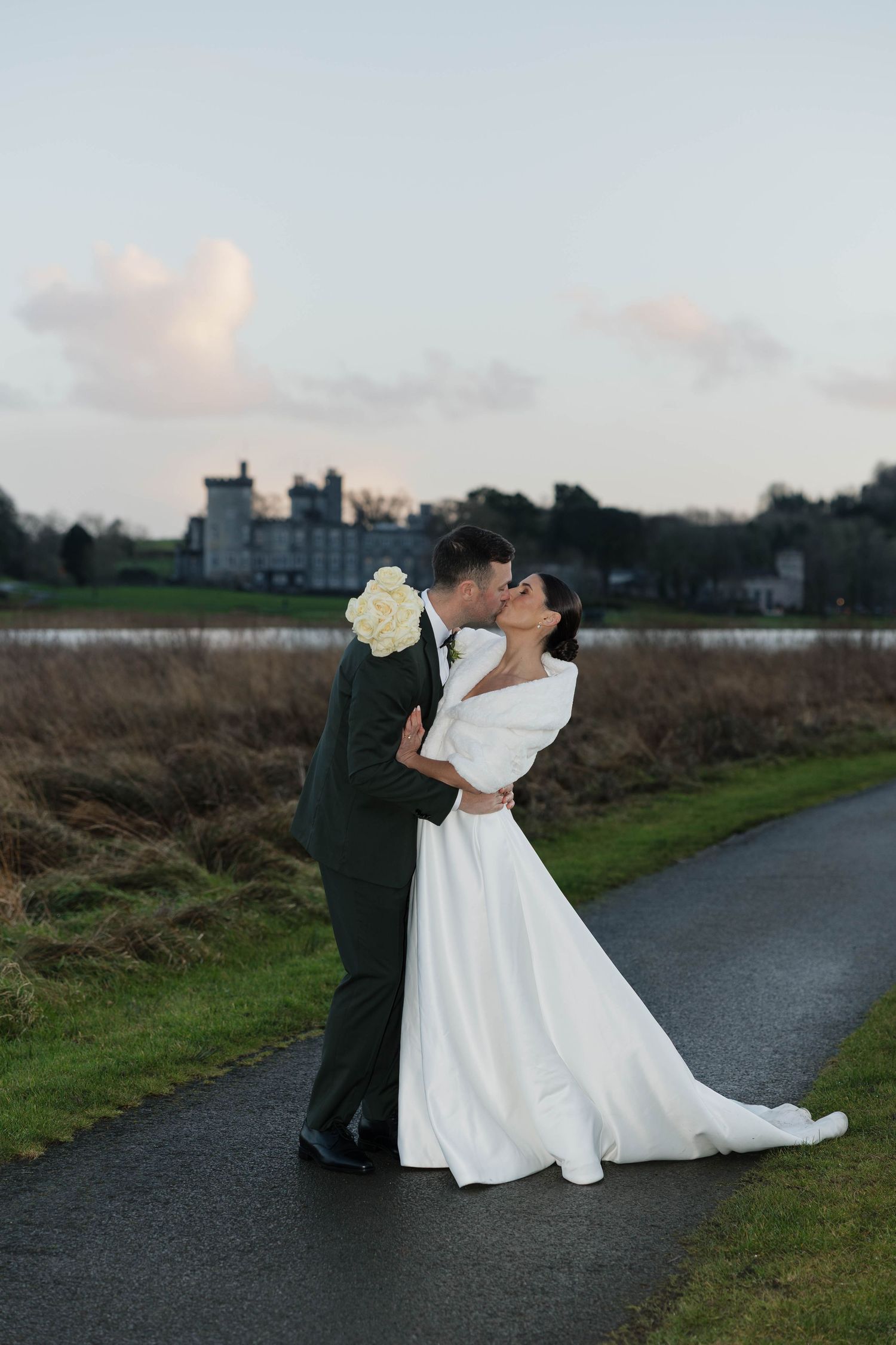 A couple in wedding attire embraces on a path with a castle in the background at dusk.