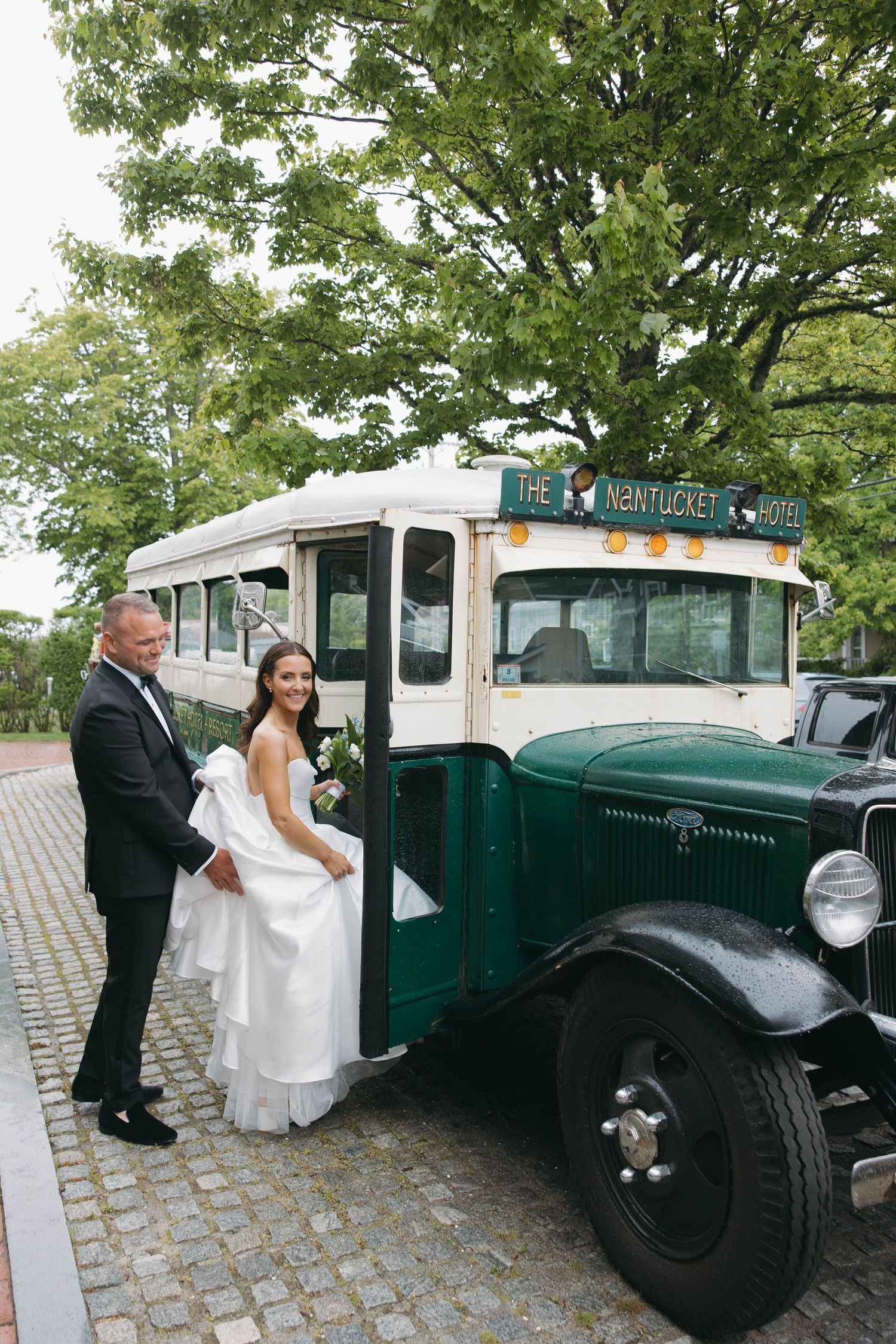 Newlyweds pose by vintage green hotel transport vehicle on cobblestone driveway.