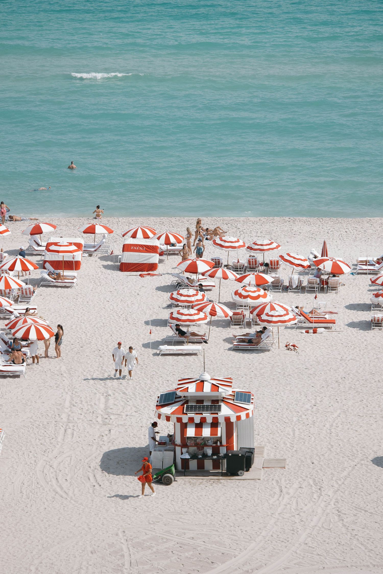 Aerial view of a beach with red and white striped umbrellas and lounge chairs on white sand.