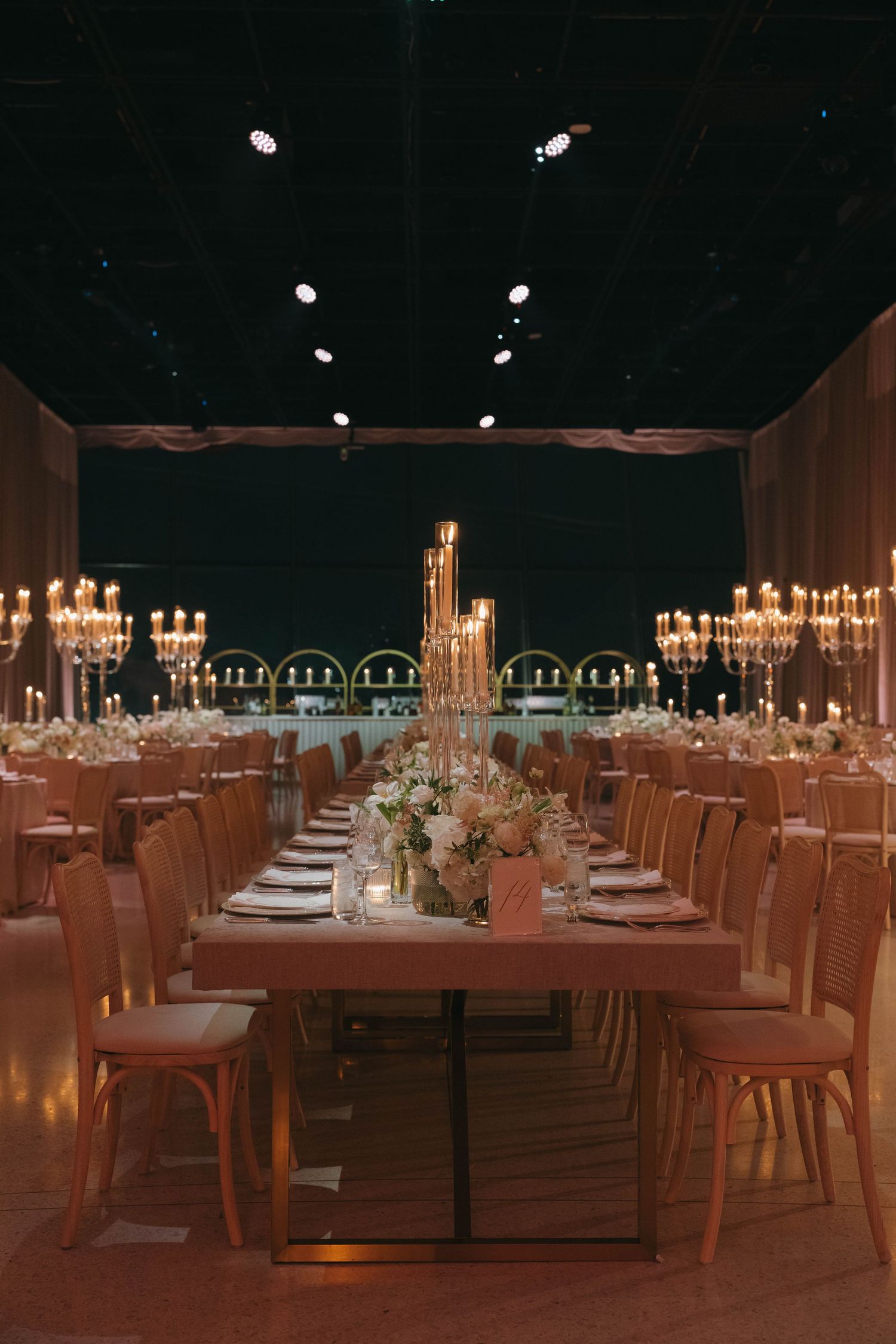 Dimly lit reception hall with long tables arranged in rows, illuminated by candlelight and chandeliers.