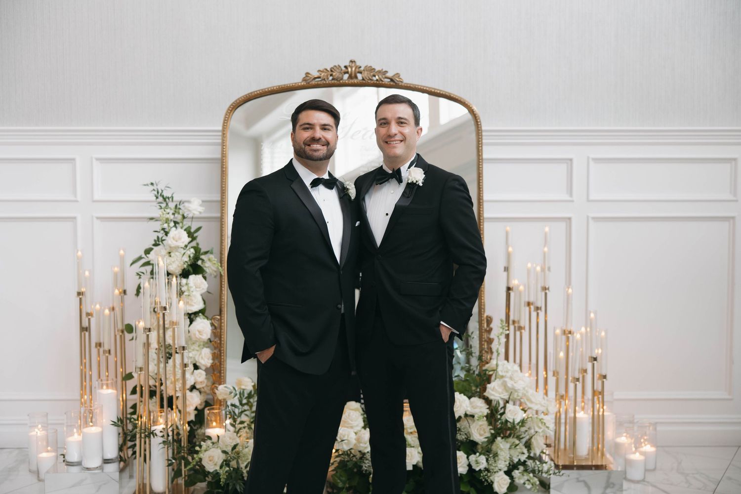 Wedding party members stand together in front of an ornate gold mirror with floral arrangements.