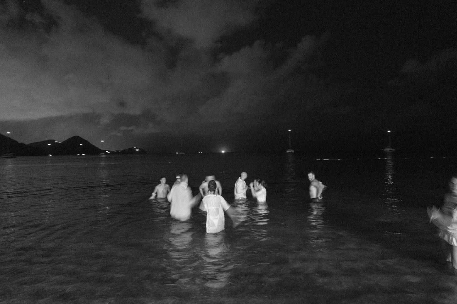 Group of people wading in ocean at night, black and white photo with distant lights on horizon.