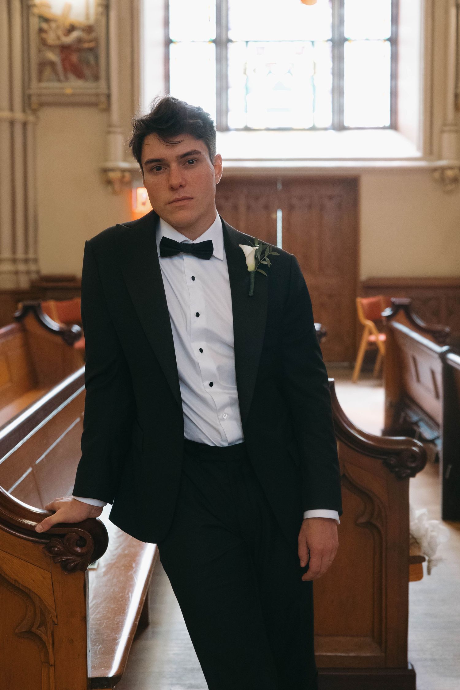 Young groom in black tuxedo with bow tie and boutonniere standing in church aisle between wooden pews.