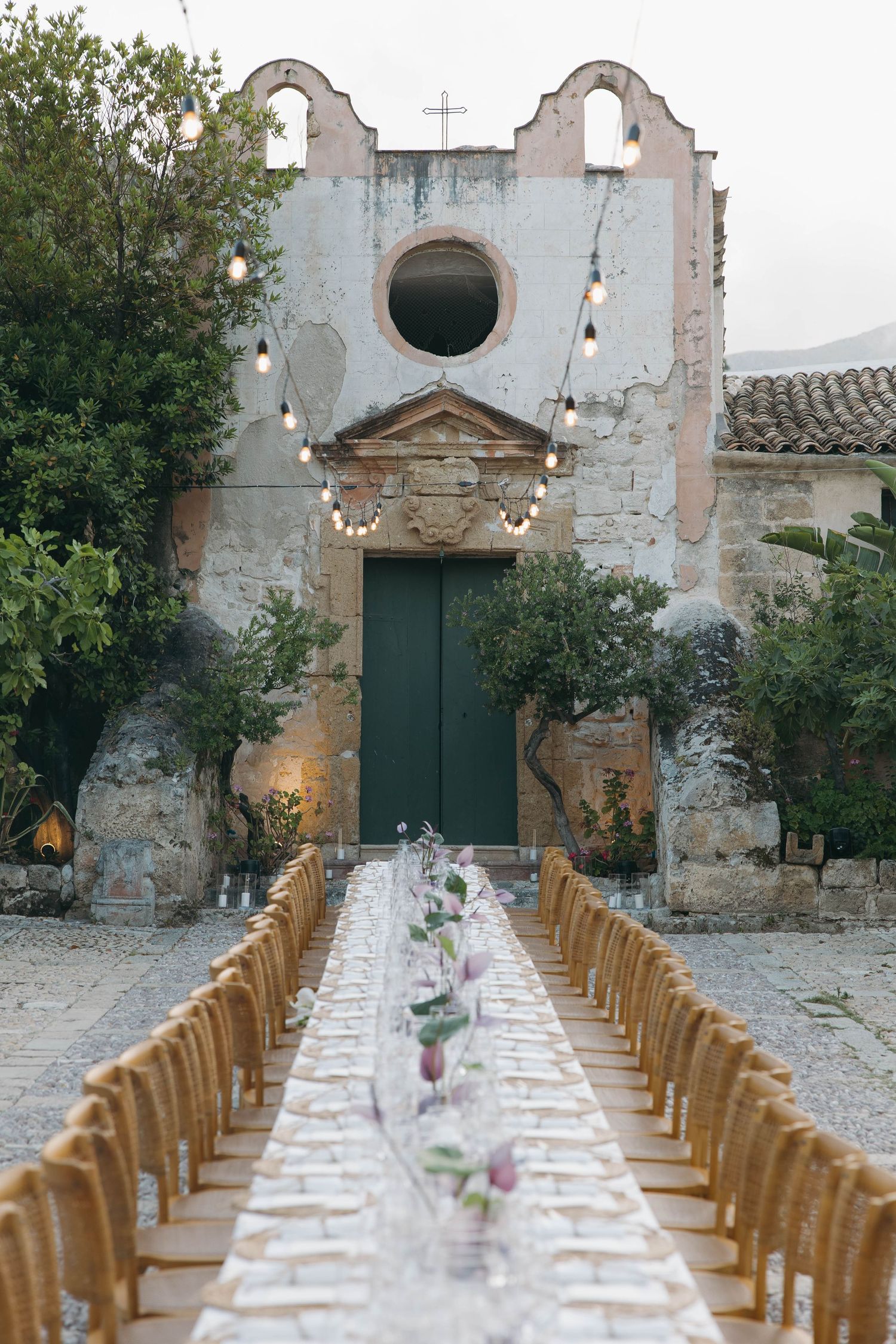 Long outdoor dining table setup with string lights and wooden chairs against historic stone building.
