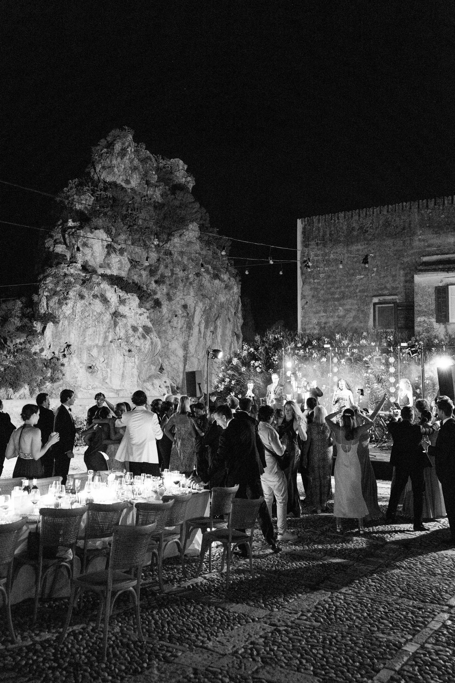 Elegant evening celebration with guests mingling around illuminated tables in a stone courtyard.