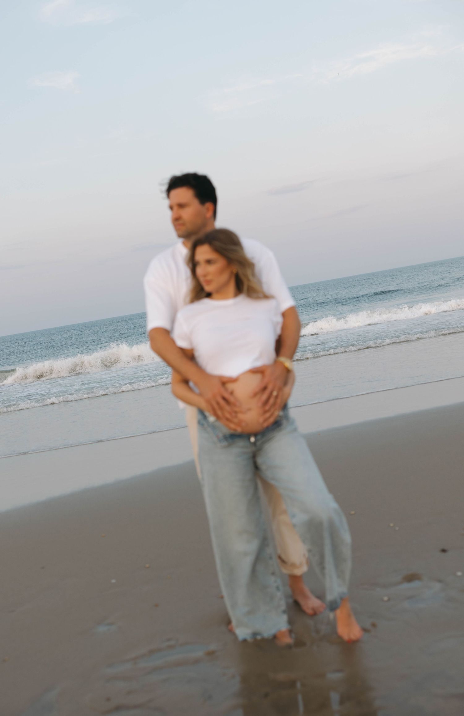 A romantic couple embraces on a serene beach at dusk, both wearing casual white tops and jeans.